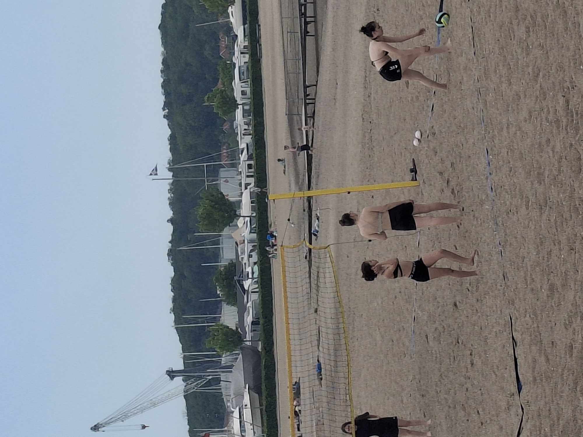The image depicts a group of people playing beach volleyball on a sandy beach in Aabenraa, Denmark. In the background are parked vehicles and boats, suggesting a marina or waterfront area. Trees line the coast behind the vehicles, and a crane is visible on the left side of the image. A beach volleyball net divides the players. In the upper right, a woman kicks a volleyball. Other players are on both sides of the net, in positions ready to play. The sky is clear.