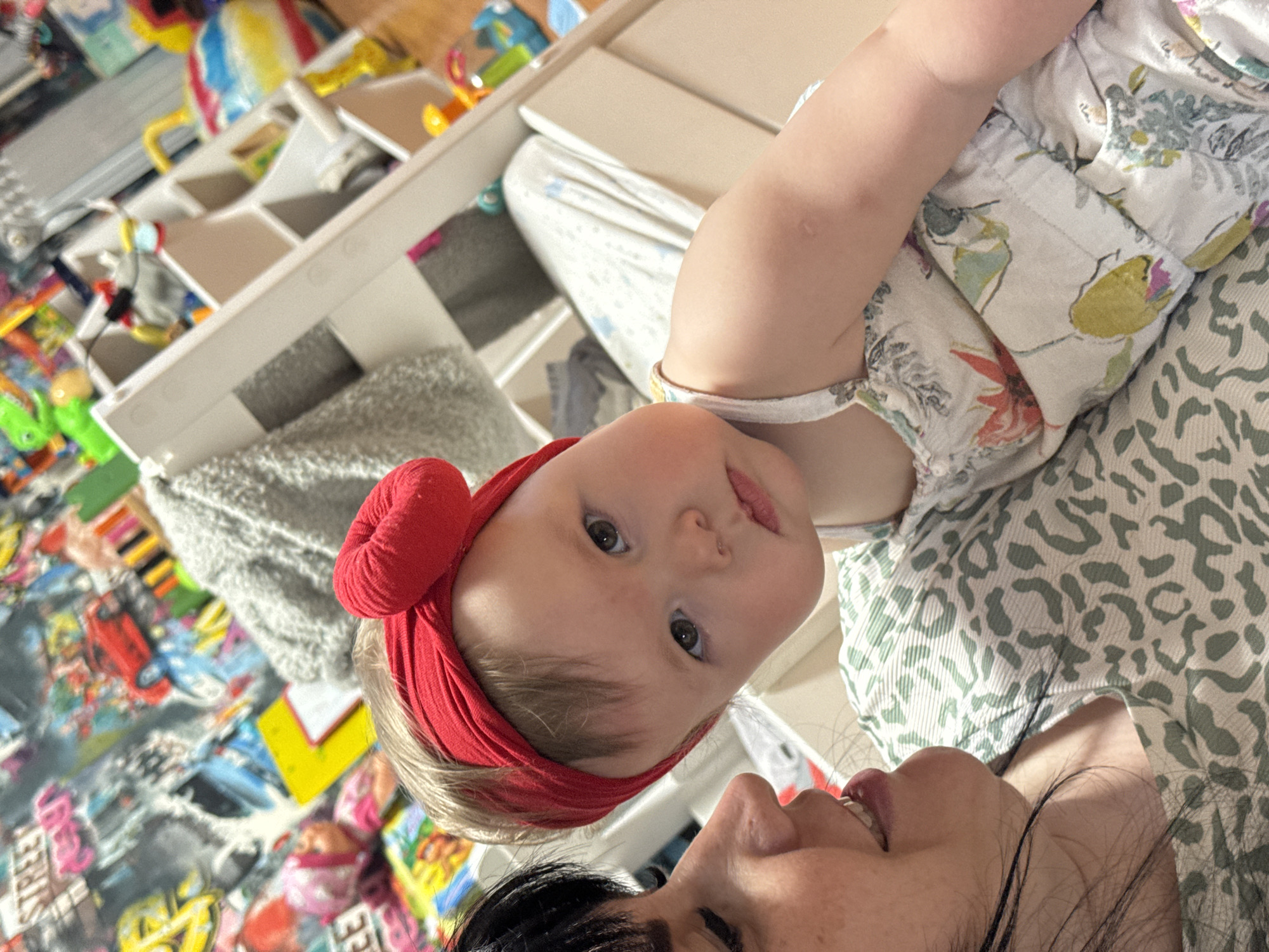 A close-up, slightly rotated, shot depicts a baby with a red headband and a woman lying down together. The baby, positioned above the woman, has fair skin, light hair, and brown eyes. The woman, situated below the baby, has dark hair. They appear to be indoors, possibly at home, given the presence of toys and shelves in the background. The environment appears cluttered with various items. The picture shows an intimate moment between the mother and her baby. The city of Zav'yalovo, Russia is not discernible from the image.