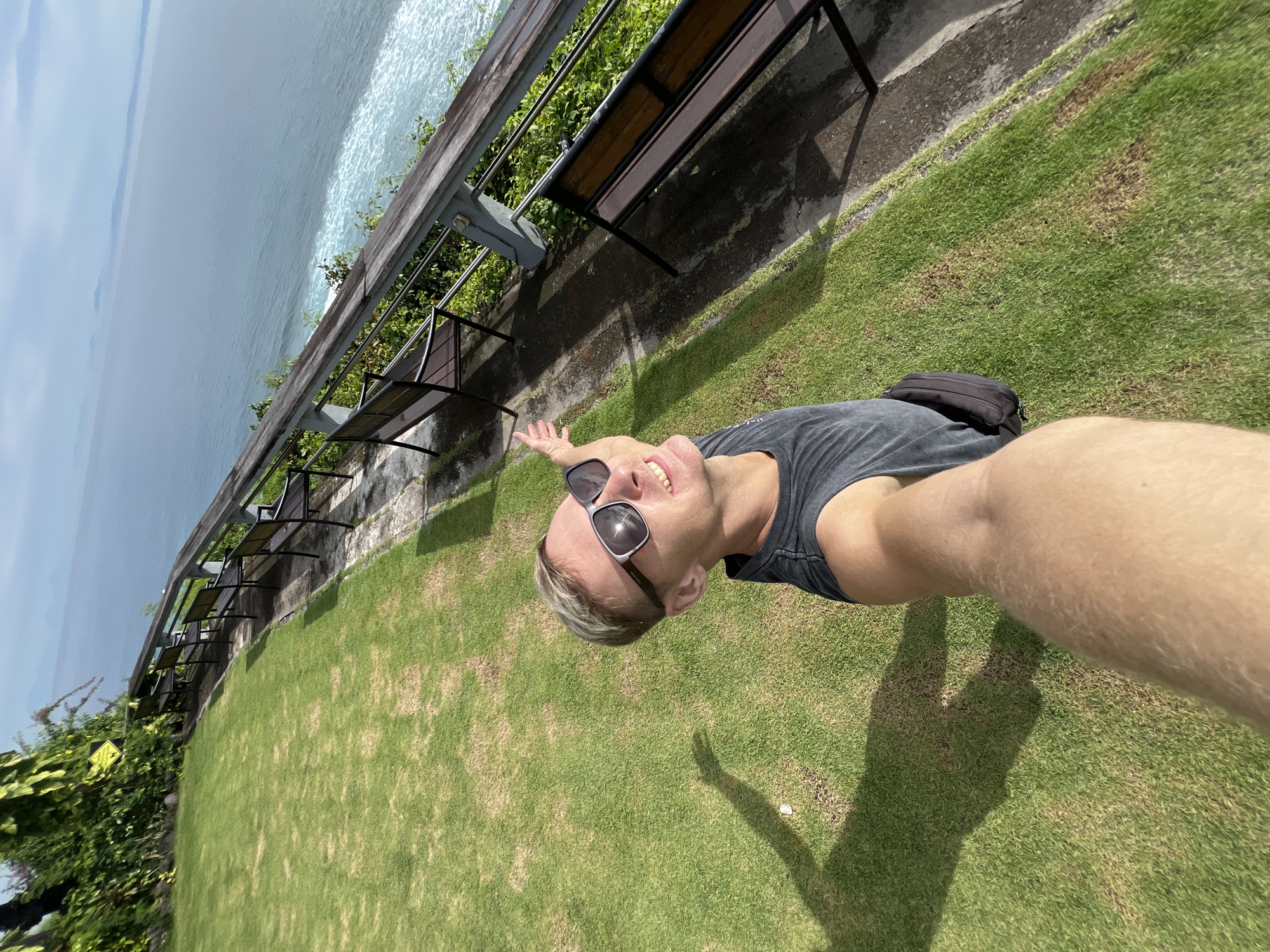 A man stands on green grass on a sunny day in Kuta Selatan, Indonesia. The man is wearing sunglasses and a dark t-shirt, with a black bag across his chest. He extends his arm towards the camera as if taking a selfie. Behind the man is a row of dark benches situated along the edge of a cliff overlooking the ocean. A metal railing runs along the cliff edge with some green plants growing at the base of it. The sea is visible in the background.