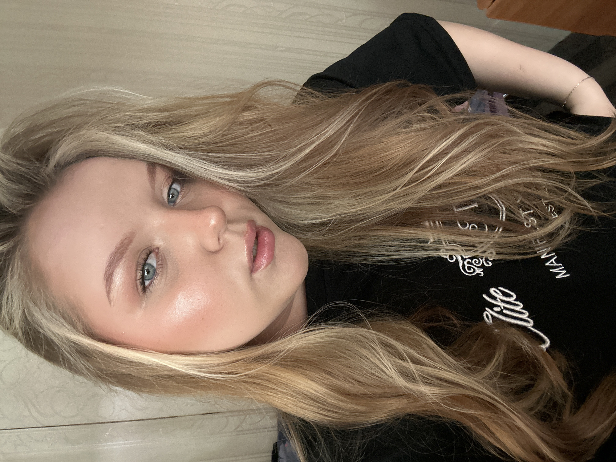 A fair-skinned woman with blonde hair lies on her side, facing the camera. She has light blue eyes and appears to be wearing light makeup, including blush and lip gloss. She is wearing a black t-shirt with white lettering that reads "life" and a partial word that looks like "manifest" on it. Her hair is long and wavy, with lighter highlights throughout. A silver bracelet is on her left wrist. The background is blurry, suggesting an indoor setting, with a cream colored door is visible. There is no evidence indicating location in the city of Noviy, Russia.