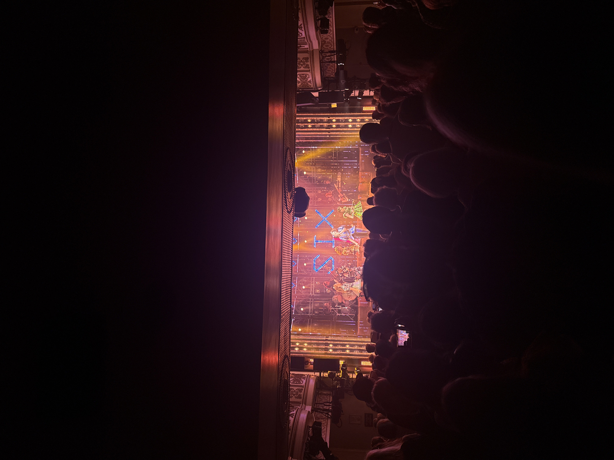 The image shows a performance of the musical "SIX" on a stage, viewed from an audience perspective. The stage lighting is prominent, with the word "SIX" spelled out in illuminated letters. Performers can be seen on stage, and the audience is visible as dark shapes in the foreground. The location is a theater in an unknown city. The picture is rotated ninety degrees.
