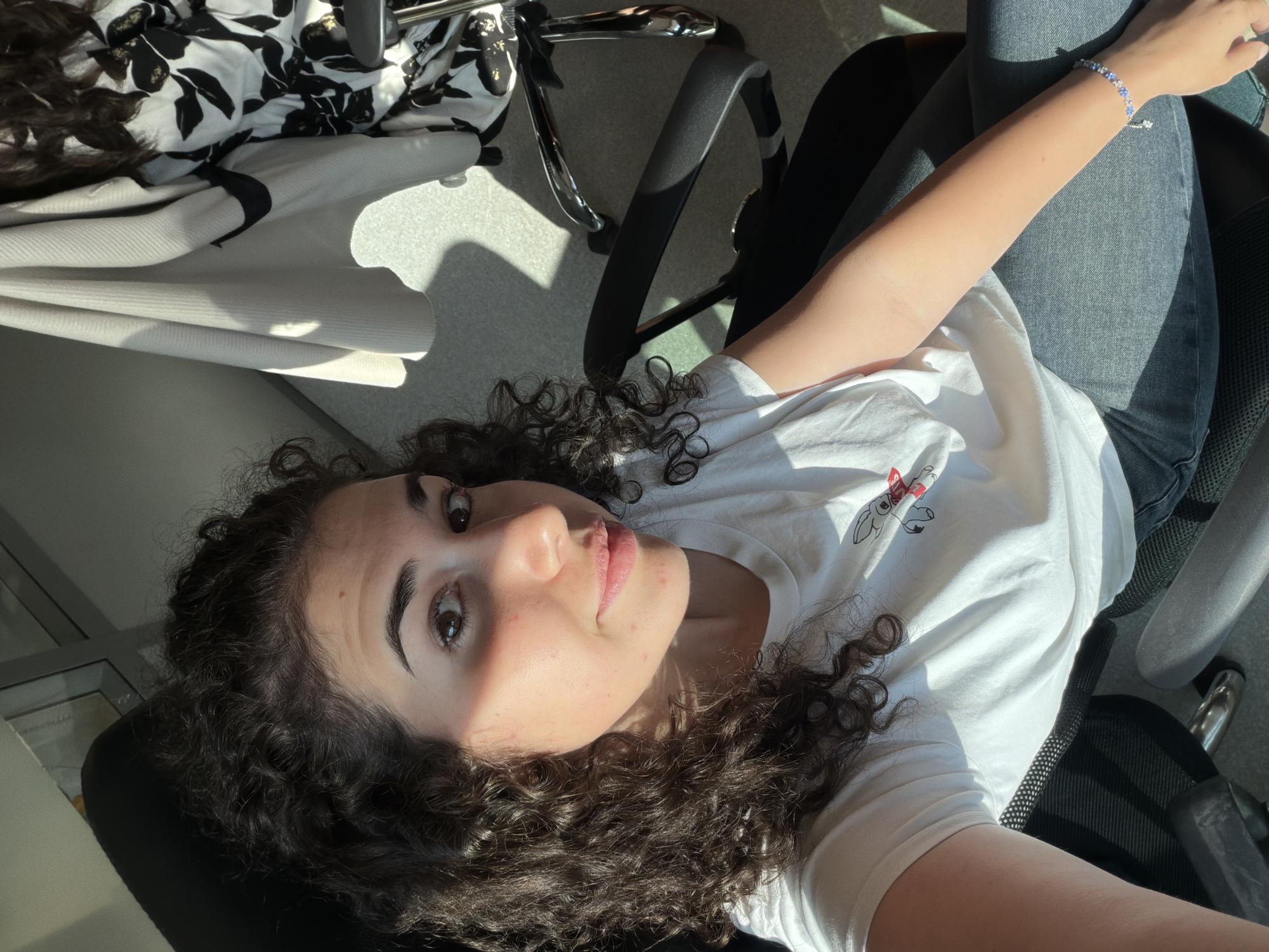 In Sofia, Bulgaria, a young woman with curly, dark brown hair sits in a black office chair, her body angled towards the camera. She is wearing a white t-shirt with a small cartoon rocket graphic on it and dark jeans. Sunlight streams in from the right, casting a distinct shadow across the scene and illuminating her face, highlighting her brown eyes and a faint smile. Her left arm rests on the armrest of the chair, and a blue beaded bracelet adorns her wrist. In the background, a white jacket is draped over another chair, and part of a black and white floral pattern is visible. The environment appears to be an indoor setting, possibly an office or studio, with a light-colored floor and wall.