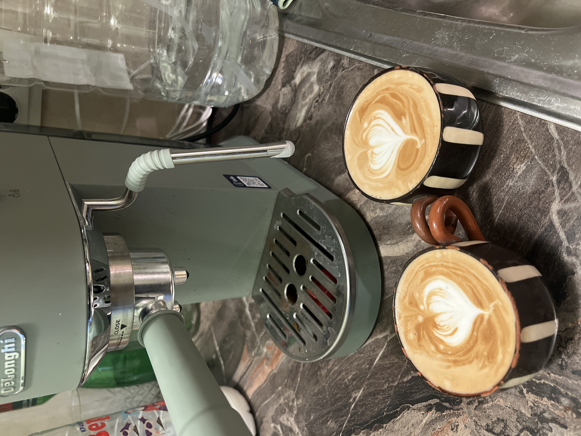 The scene depicts a kitchen countertop with a DeLonghi espresso machine on the left and two cups of latte art coffee on the right. The espresso machine is light green with chrome accents. A clear plastic water container sits behind it. The coffee cups are black and white striped with heart-shaped latte art. The countertop has a dark, marble-like pattern. No people are visible. There is no information to suggest the location is in Plovdiv, Bulgaria.