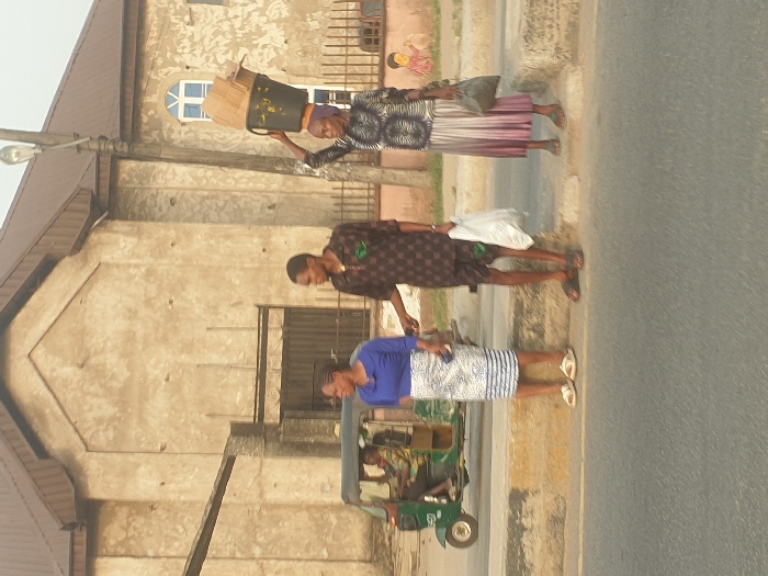 Three people walk along a road's edge next to a building. One woman carries items on her head. A man in a patterned shirt walks between her and another woman in a blue top. A three-wheeled vehicle is nearby. The building behind them is a light-colored, two-story structure with a brown roof, bars on the windows, and a faded mural on an upper wall. The road appears to be asphalt.