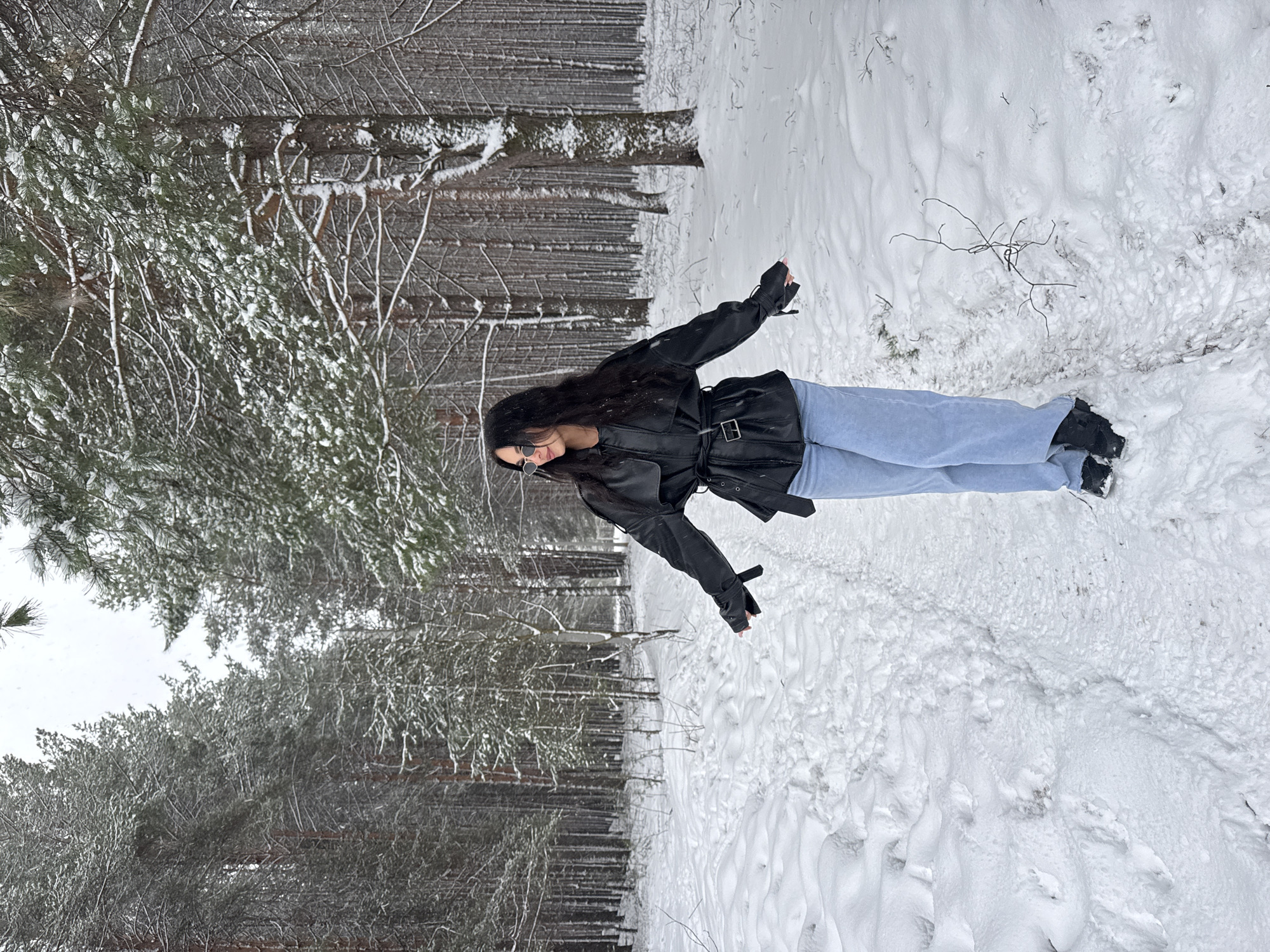 A person is lying on their back in the snow, arms outstretched, mimicking the form of a snow angel. They are dressed in light blue jeans and a black leather jacket with a belt. Sunglasses are worn on their face. The location is a snow-covered forest, possibly on the outskirts of the city of Chaikovsky, Russia. The trees are partially covered in snow, and the ground is entirely blanketed in white. The scene conveys a sense of winter recreation in a forested environment.