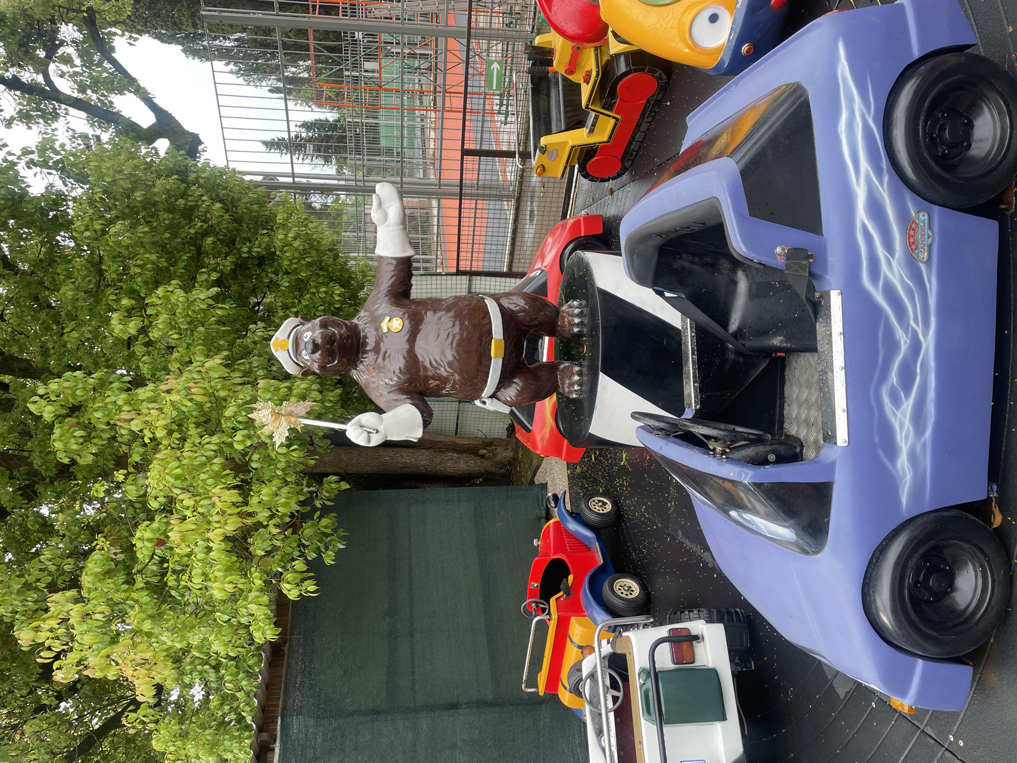The image shows a children's amusement ride in Sochi, Russia. A brown bear statue with a white policeman's hat and gloves sits in the driver's seat of a blue car with white lightning bolt designs. The car is surrounded by other kiddie rides, including a red and yellow car, a yellow bulldozer, and a white vehicle with a green seat. In the background, there's a green leafy tree, a fence, and what appears to be a sports court or playground area. The scene suggests an outdoor amusement area or park.