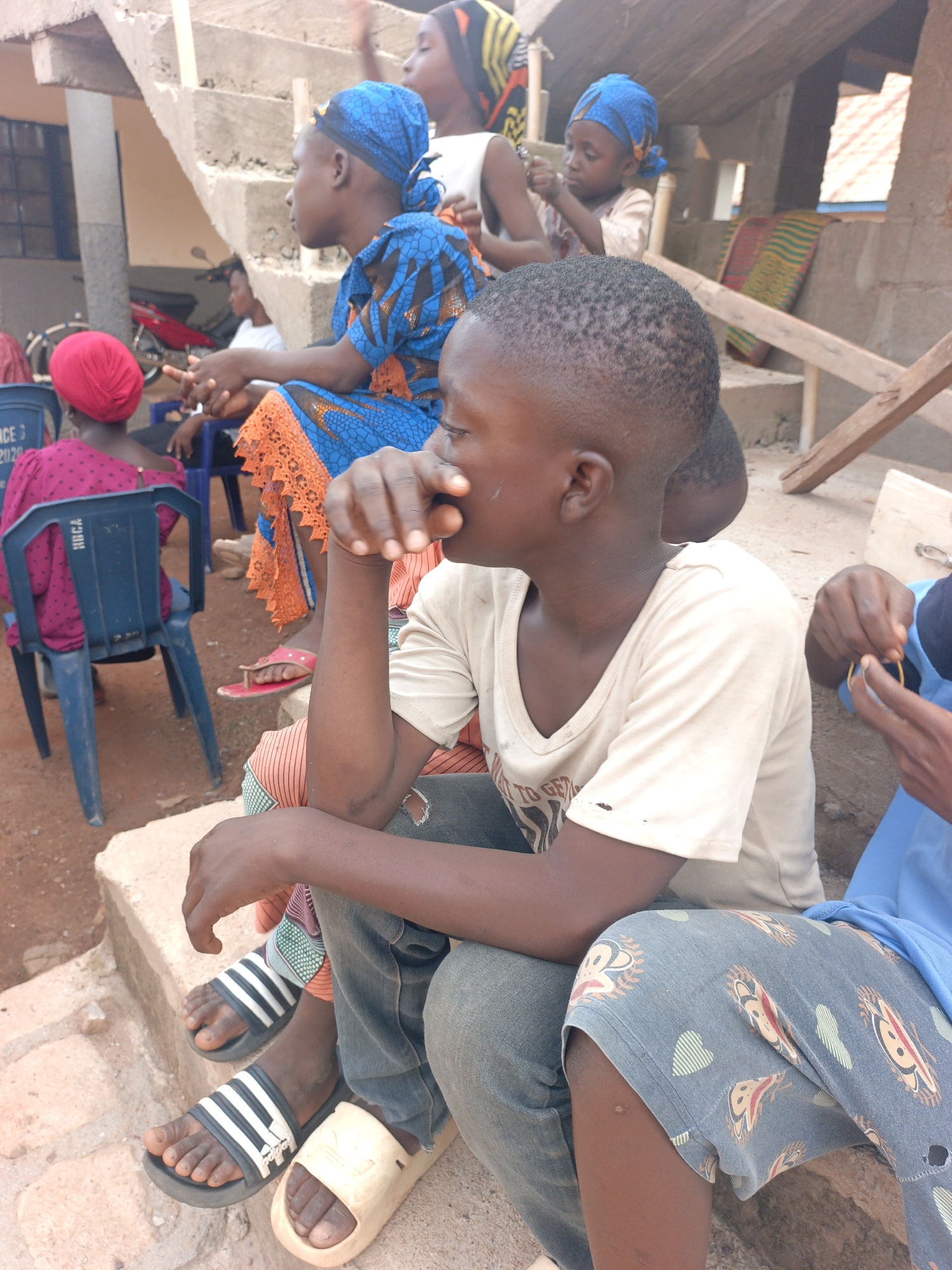 The image captures an outdoor gathering of several individuals, primarily children and young adults, in Akwanga, Nigeria. The scene is set around concrete steps, suggesting a residential or community area.
In the immediate foreground, a young boy with closely cropped hair is seated on the steps, wearing a light-colored V-neck t-shirt and blue jeans, with one hand covering his mouth. He is looking towards the right side of the frame. Next to him, another person is partially visible, wearing blue shorts with a pattern of hearts and cartoon-like faces, and holding a yellow circular object or band in their hands.
Further back, a young woman or older girl is seated on the steps, dressed in a vibrant blue and orange traditional patterned garment with a matching blue headwrap. Her red flip-flops are visible below. Behind her, another individual wearing a red headwrap is seated on a blue plastic chair. The back of this chair displays partially visible text: "ICE 3 2020" and possibly "HRCA". A small child in a blue headwrap is also seen higher up on the steps.
Other people are present in the mid-ground and background, some partially obscured. A red motorcycle is visible behind the chairs. The overall setting includes simple concrete structures and what appears to be a building in the background. The bright lighting indicates it is daytime. The people appear to be casually seated, observing their surroundings or interacting quietly.