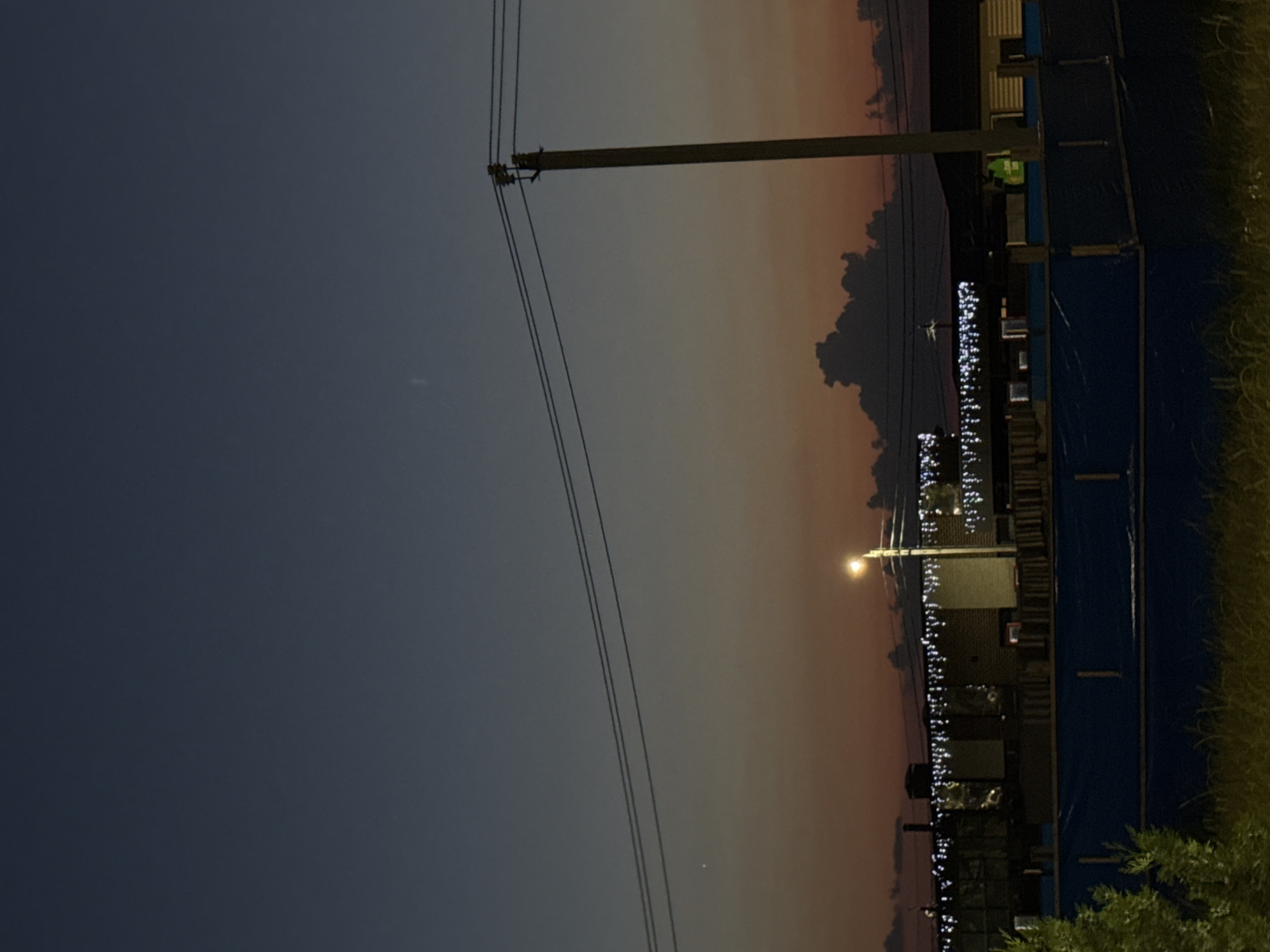 The image depicts a nighttime scene in Mikhaylovsk, Russia, possibly during twilight due to the soft orange glow in the sky. A power line pole with wires stretches across the frame, casting a shadow. In the background, a building or structure is visible, adorned with numerous small, bright lights, likely decorative. A street lamp illuminates the foreground to the right, its light casting a warm glow. The setting appears to be a somewhat urban or suburban area, with hints of grass and foliage to the far right. The overall mood is quiet and serene, typical of a late evening. There are no people visible in the frame.