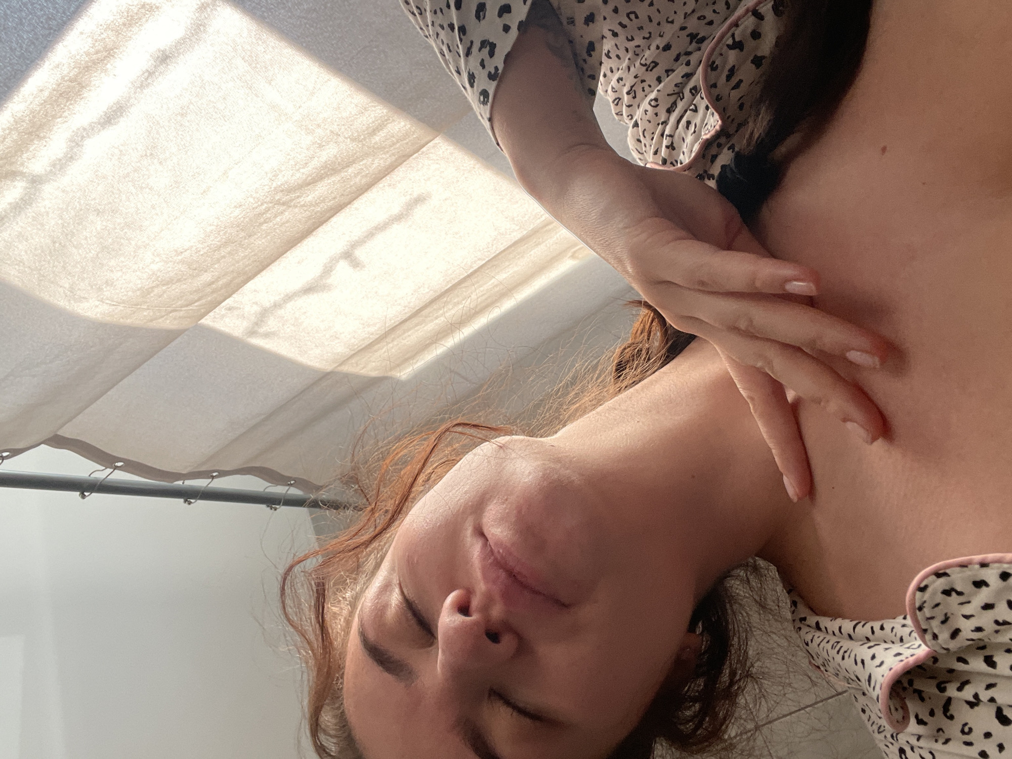 A woman is lying down, likely indoors, with her eyes closed. She is wearing a spotted pajama top with a pink trim. Her right hand rests on her upper chest/shoulder area. Part of a window with sheer white curtains is visible in the background. The image has been rotated counterclockwise. There is no evidence indicating this photo was taken in Мосметаллоконструкция Онт, Russia.