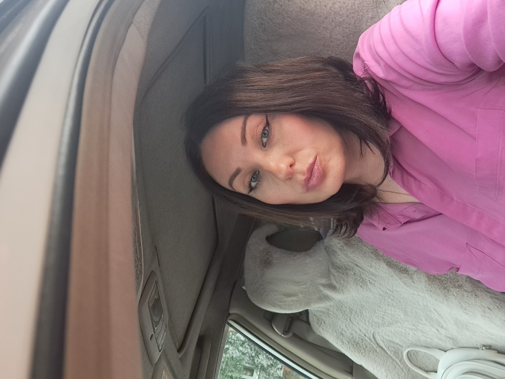 A woman with dark brown hair and light green eyes, wearing a pink button-up shirt, takes a selfie from the driver's seat of a car. She has winged eyeliner and pink lipstick on. To her left, a car's interior is visible, including a fuzzy, light-colored seat cover on the passenger seat and a white handbag resting on it. The car's ceiling, with an overhead light fixture, is also in view. Outside the car window, trees and a building can be seen, suggesting an outdoor setting, potentially in a residential area. The lighting indicates it is daytime. This image was captured in Irkutsk, Russia.