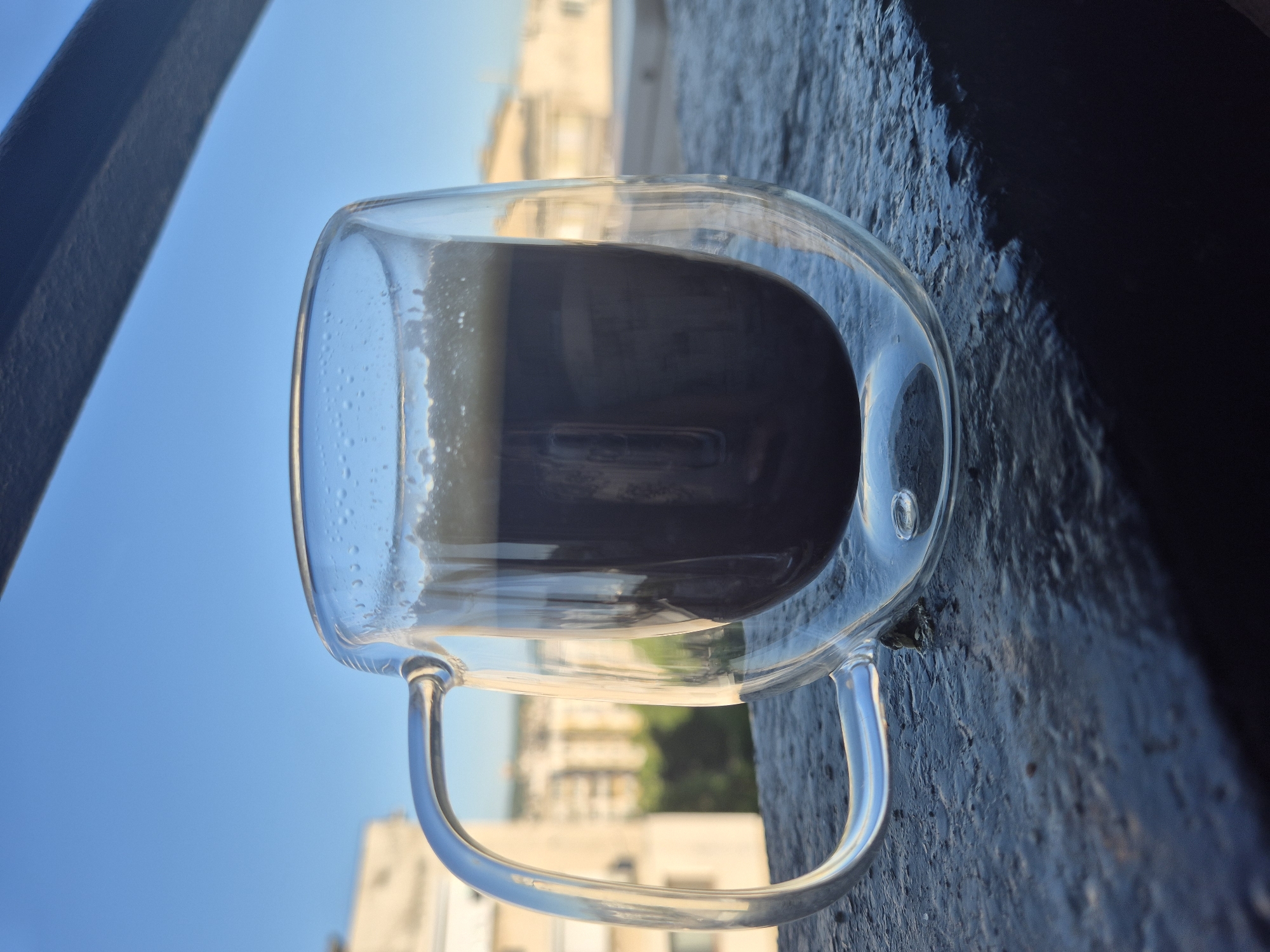 A clear glass mug filled with a dark liquid rests on a dark-colored, possibly textured, surface. The mug has a handle and a double-walled design. The dark liquid inside appears to be coffee or a similar beverage. In the background, the scene transitions from a clear blue sky to buildings in an urban area. The buildings appear to be light-colored. A portion of a dark railing or structure is visible on the left edge of the frame.