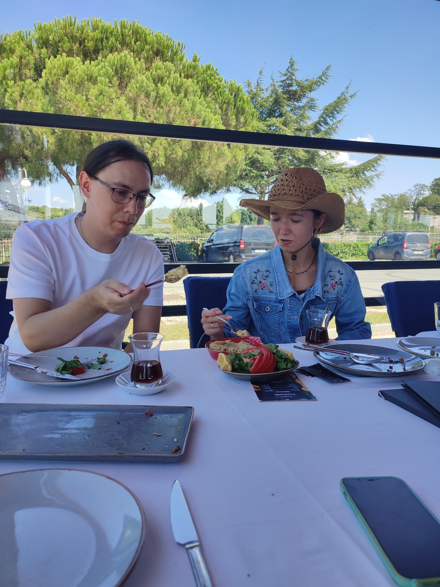 In Üsküdar, Turkey, two individuals are seated at a table covered with a white tablecloth, engaged in eating. The man on the left, wearing glasses and a white t-shirt, is holding a spoon with food, looking down at his plate. The woman on the right, sporting a straw cowboy hat and a denim jacket adorned with embroidered flowers, is also eating. In front of them are plates with remnants of food, cutlery, and small glasses of dark liquid, likely tea. A partially eaten salad with tomato and lemon wedges is visible on a plate. The scene appears to be an outdoor dining area, possibly a restaurant, with large windows offering a view of lush green trees and vehicles passing by outside. The sky is a clear blue.