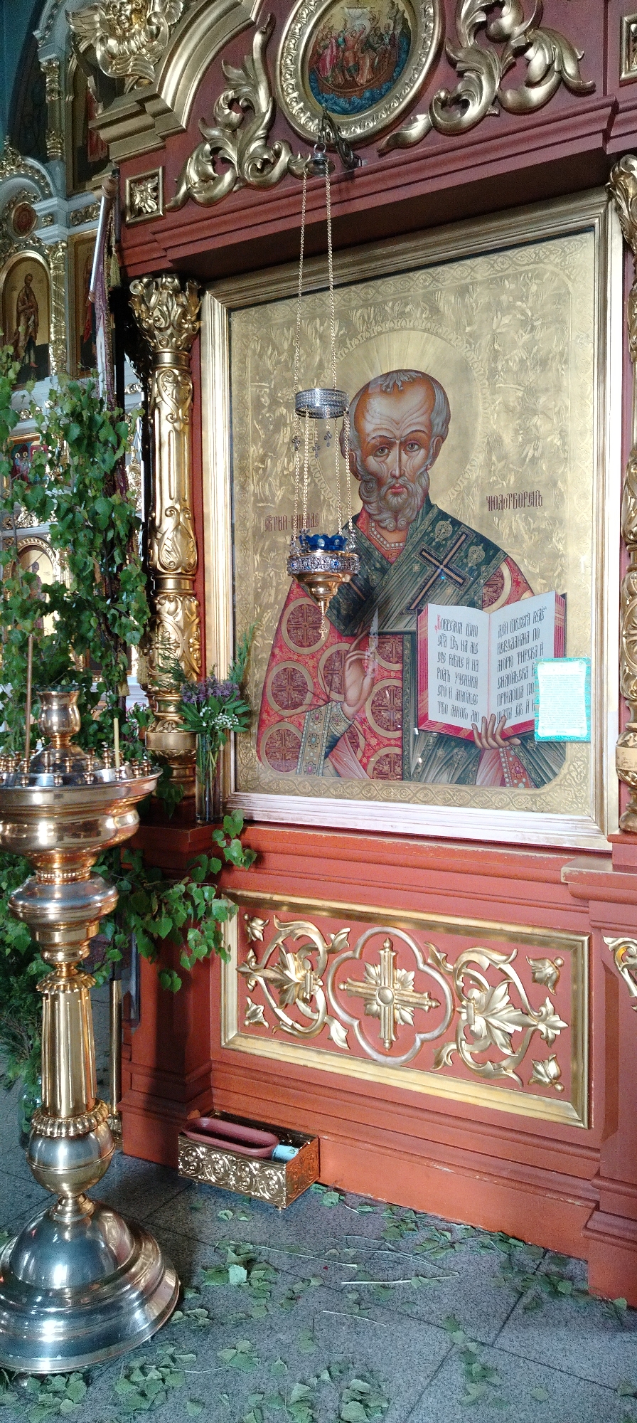 This image captures a section of a richly adorned Orthodox church interior, likely in Chaikovsky, Russia. The central subject is a large, gilded icon depicting a male saint, identified by Cyrillic text above his halo as likely "Святитель Николай" (Saint Nicholas). The saint has a white beard, a receding hairline, and a halo, and is clad in ornate red and green clerical vestments. He holds an open book, presumed to be the Gospel, in his left hand, displaying Cyrillic script within its pages, with discernible words like "УЧИМЪ" (we learn/teach) and "Слово" (Word). A small blue-bordered sticker with text is affixed to the open book.
A silver and gold vigil lamp, suspended by delicate chains, hangs in front of the icon, casting a gentle glow. The icon is framed by an elaborate golden wooden structure, which is part of a larger devotional stand or iconostasis. Below the icon, a dark red panel features intricate golden carvings, including a stylized cross within a quatrefoil design and floral patterns.
To the left of the icon, a tall, golden candelabra or lampada stand is prominent, decorated with fresh green birch branches. This adornment, along with scattered dried green leaves on the grey tiled floor, suggests a religious celebration, possibly Pentecost (Trinity Sunday), for which birch branches are traditionally used to decorate churches. More green foliage extends from behind the candelabra and around the icon. In the lower right, a small golden box holding candles rests on the floor. The scene evokes a feeling of reverence and highlights the rich visual traditions of Orthodox worship.