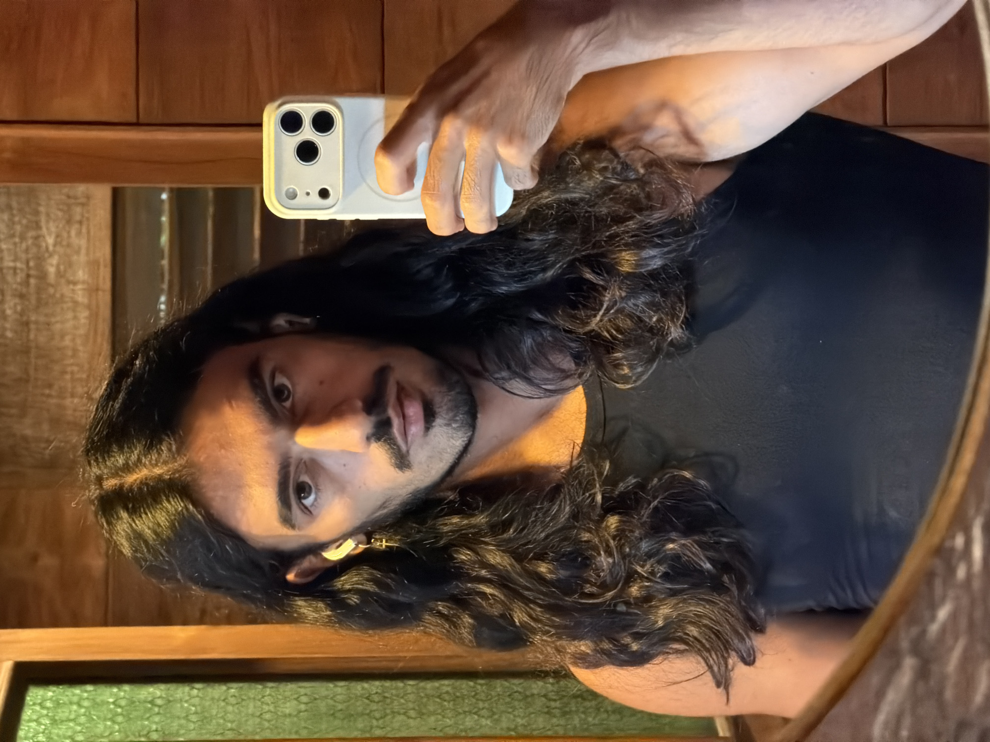 A dark-haired man with a short beard and mustache is looking at the camera in a mirror. He is holding a cell phone in his left hand. He is wearing a black tank top. The background appears to be a wooden wall, and the mirror frame appears to be wooden with a green textured area underneath the mirror, possibly reflecting a surface. The photo was likely taken in North Kuta, Indonesia, since the location was provided.