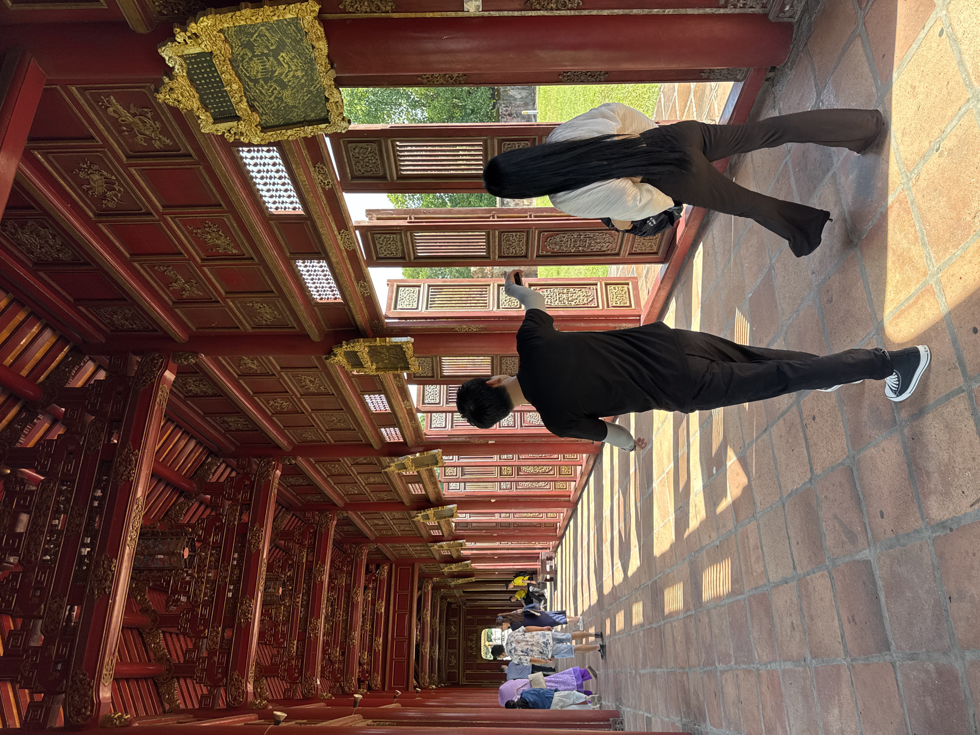 In Phú Xuân Ward, Vietnam, a long, red, covered walkway with intricate wooden and golden ornamentation is the setting. Sunlight streams through open doorways, casting striped shadows on the terracotta tiled floor. Two individuals, a man in a black t-shirt and black pants and a woman with long black hair wearing a white blouse and dark pants, walk away from the camera towards an open doorway leading to a green outdoor area. Other visitors are visible further down the walkway, walking in the same direction. The architecture suggests a historical or cultural site, possibly a temple or palace, characterized by its ornate wooden beams, panels, and decorative gold accents. The atmosphere is bright and airy, with the sunlight creating a dynamic interplay of light and shadow.
