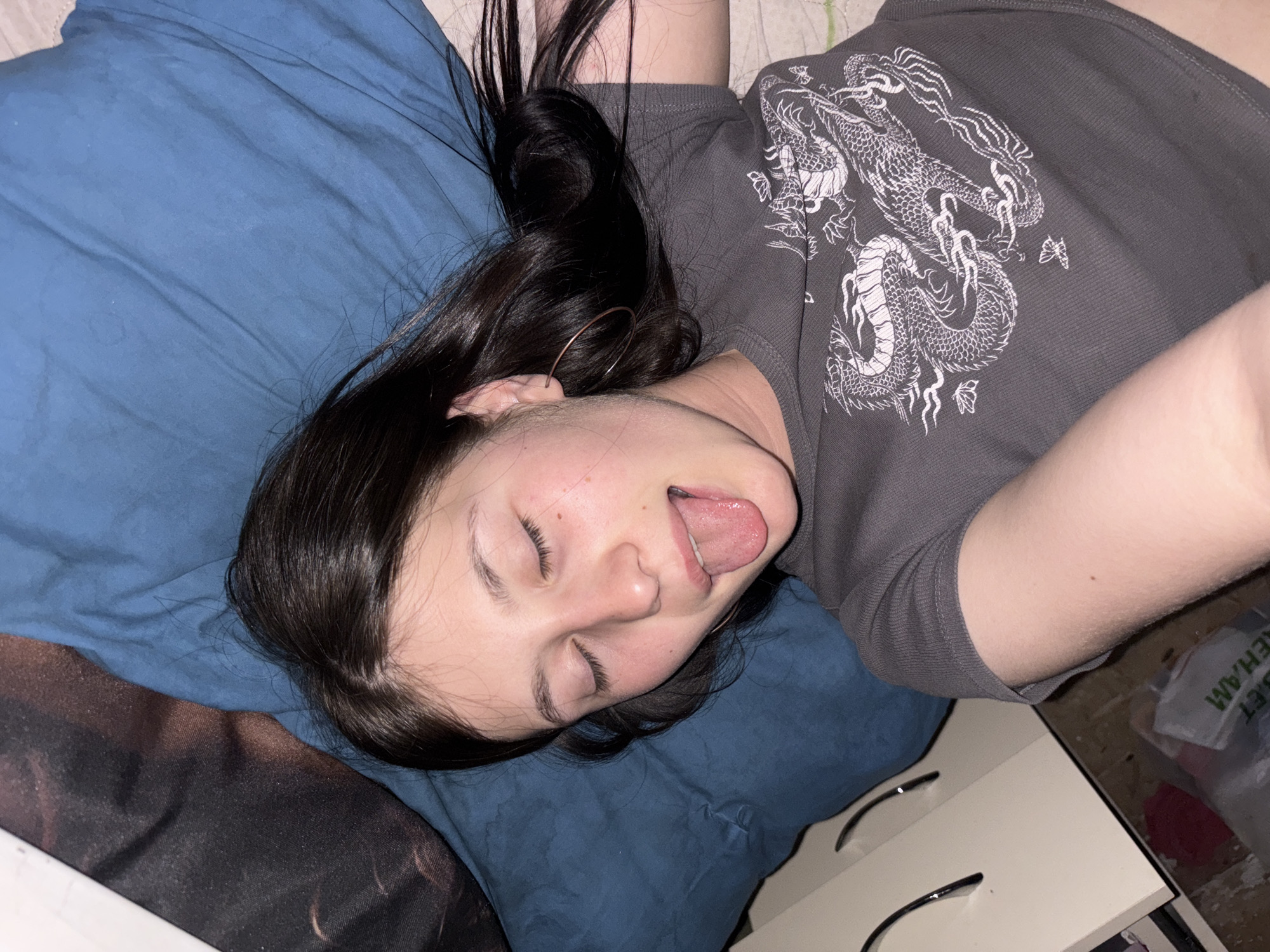 A woman is lying on a blue pillow with dark hair. She is wearing a gray shirt with a dragon design. Her eyes are closed and her tongue is sticking out. A white bedside table is visible with items on and around it. The location is not discernible from the image.