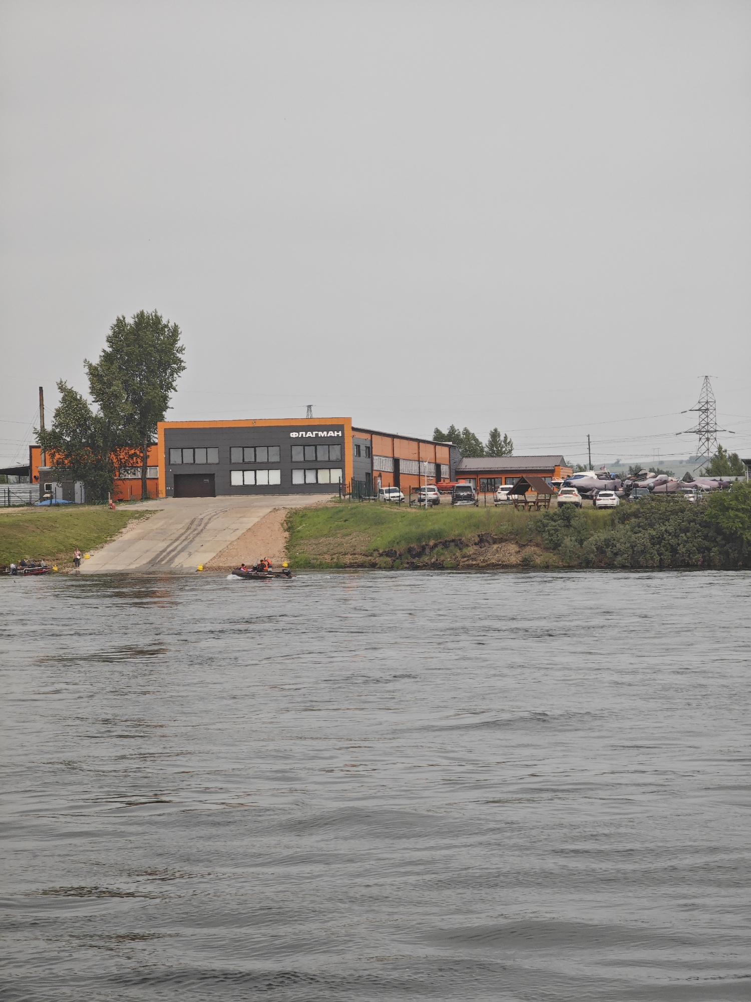 A scene unfolds on the bank of a body of water, likely a river, on an overcast day. A modern, multi-story building with orange and dark grey sections dominates the middle ground. The building features large windows and a prominent entrance with a ramp leading down to the water. The signage on the building, in Cyrillic letters, reads "ФЛАГМАН".
To the left of the building, a tall, slender smokestack rises into the sky, and several trees are present. To the right, a high-voltage power line tower is visible in the distance. The bank of the river is covered with green grass and some low-lying bushes.
In the water, two boats are in motion. One, a dark-colored inflatable boat with several people wearing orange vests, is moving from left to right, creating ripples on the water. Another boat, a red one with people, is further to the left, closer to the bank. On the ramp and the bank, several individuals are observed, some appearing to be near the boats or the building. Several cars are parked on the grassy area next to the building, and a small wooden gazebo is also present. The overall environment suggests a functional area, possibly a marina or a boat service center, located in the city of Peschan, Russia.