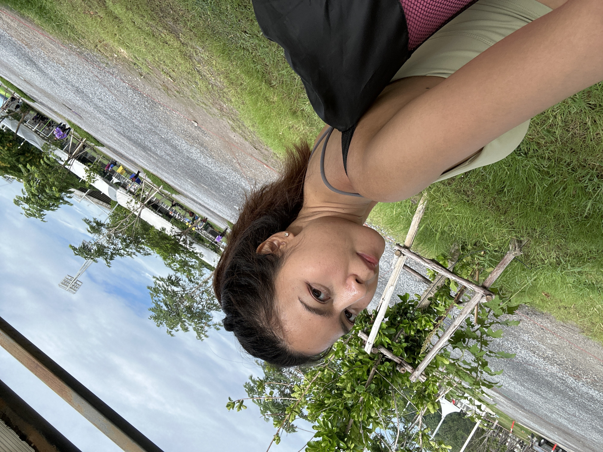 A woman is pictured in a selfie-style shot outdoors, possibly in a park or recreational area within Tha Khlong Town Municipality, Thailand. She is looking towards the camera with a neutral expression, her hair pulled back in a ponytail. She is wearing a light-colored tank top and has a black bag draped over her shoulder. The background shows a gravelly area with scattered trees, grass, and a cloudy sky. In the distance, there are tents and what appear to be stadium lights, suggesting the location might be near a sports or event venue.