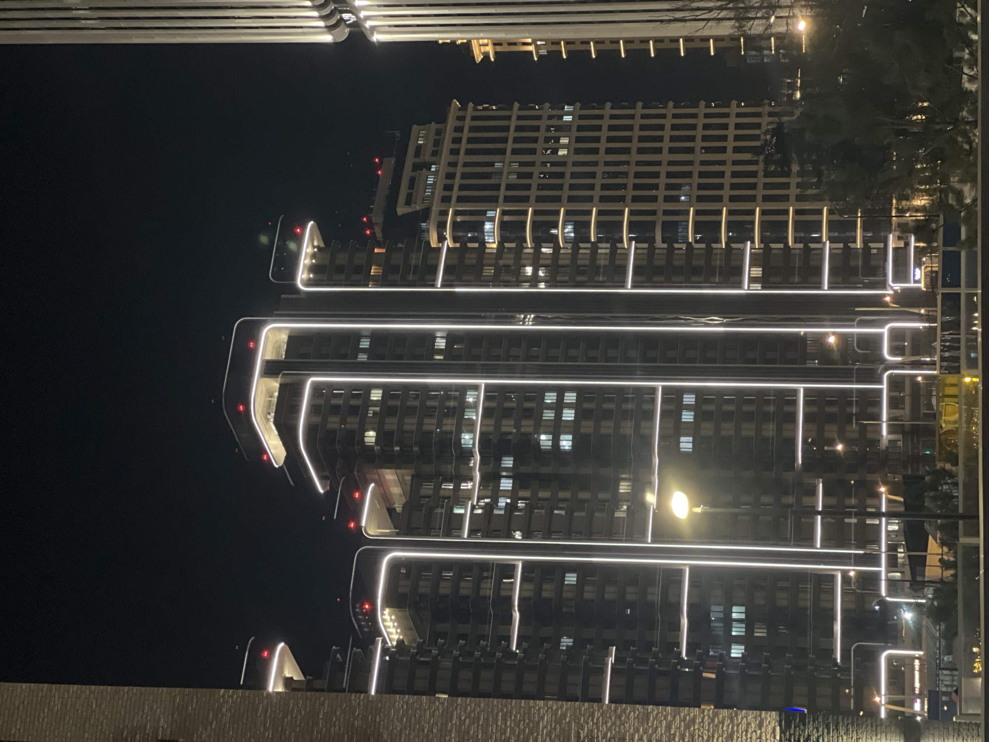 At night, the image captures a view of a modern building in Moscow, Russia, illuminated with bright white lights tracing its architectural lines. The building is tall, with numerous windows visible against the dark sky. A row of lights is horizontally oriented on the top of the building. The image orientation is rotated such that the buildings appear to be vertically stacked, rather than upright. In the foreground, the top of another structure is visible. Trees obscure portions of the view.