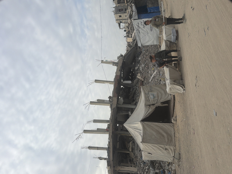 An outdoor scene in Jabalia, Palestinian Territory, depicts an area of extensive structural damage under an overcast sky. In the foreground, an unpaved, light brown dirt or sand surface extends.
In the midground, a light-colored canvas tent, bearing a blue circular logo (likely indicating a humanitarian organization), stands with its entrance partially open. To the right of this tent, three young males are positioned. Two boys stand close together, partially obscured by a low, rectangular object covered with light fabric. A third boy stands further right, facing the camera, holding a light-colored pole horizontally.
The background is dominated by the severely damaged remains of a multi-story concrete building, collapsed into a large pile of rubble. Exposed concrete columns with protruding rebar stand vertically from the debris. Further to the right in the background, additional damaged structures and another light-colored temporary tent, displaying Arabic text and numbers, are visible. The overall environment indicates significant destruction and makeshift living arrangements.