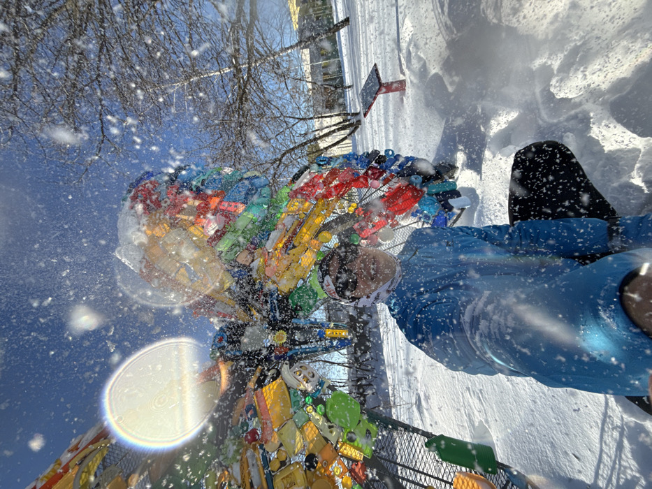The image captures a winter scene at Uram Extreme Park in Kazan, Russia, under a clear blue sky with visible airborne snowflakes or water droplets. A single individual is positioned in the lower-left foreground, facing the camera in a crouched or kneeling posture. This person wears a bright blue long-sleeved jacket, dark trousers, dark sunglasses, and a white and red patterned headband. Behind the individual stands a large, multi-colored sculpture shaped like a butterfly, constructed from numerous recycled plastic bottles and plastic components attached to a metallic framework. The sculpture's upper surfaces are dusted with snow. The ground across the scene is uniformly covered in thick snow. In the background, numerous bare deciduous trees, also covered with a light layer of snow, are visible, with distant light-colored buildings partially discernible through them. To the right of the butterfly sculpture, a red pole topped with a small, dark, angled solar panel is present. A prominent circular lens flare, indicative of a bright sun, is visible in the upper-left quadrant of the image.