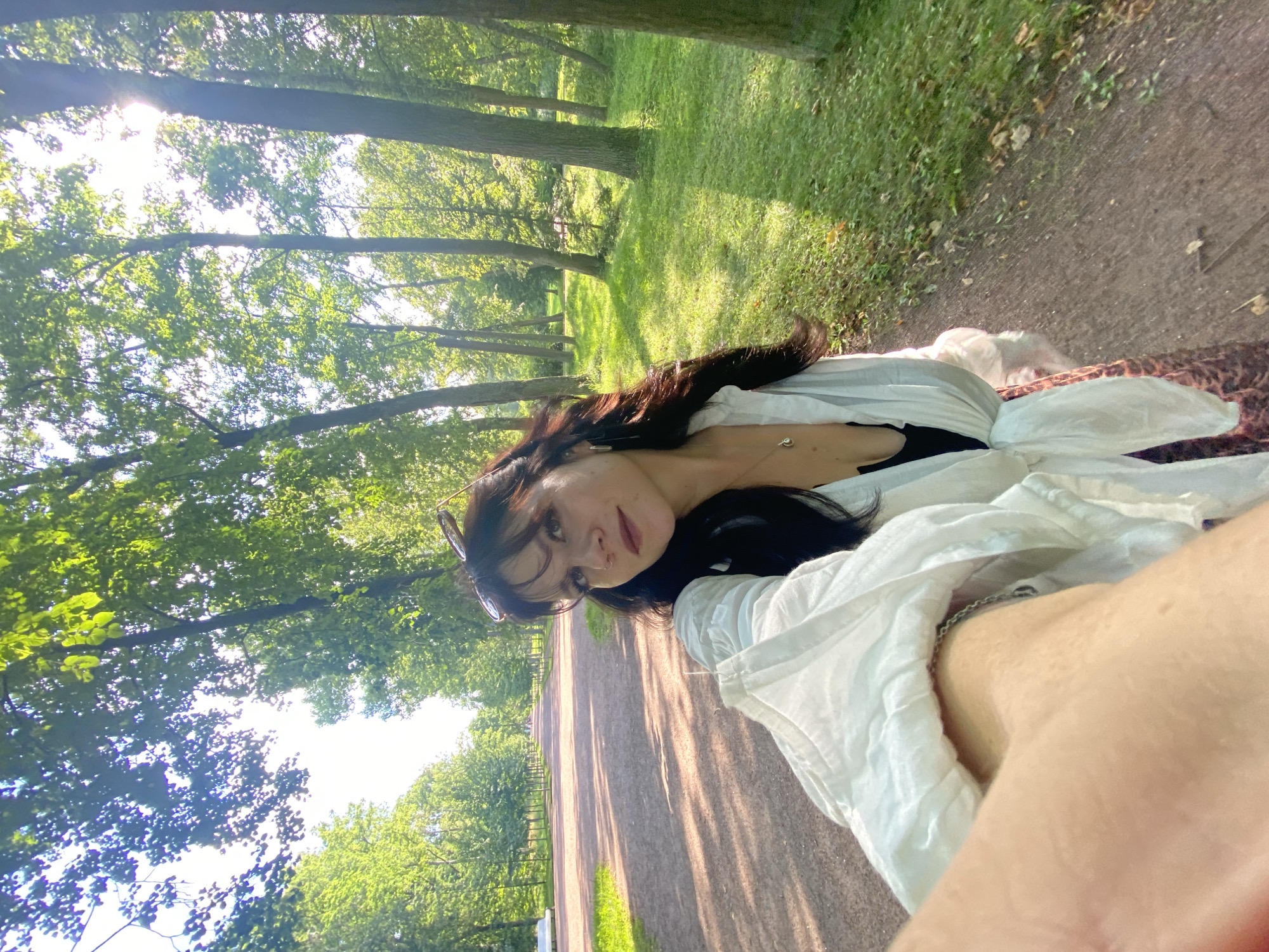 A young woman with dark, wavy hair and sunglasses perched on her head is taking a selfie in a park. She is wearing a white open blouse tied at the waist over a black top, and a patterned skirt. She is standing on a dirt path lined with trees. To her left, the path extends into the distance, flanked by a manicured lawn and more trees. Sunlight filters through the leaves, casting dappled shadows on the ground. The location is the park at the Grand Menshikov Palace in Lomonosov, Russia.