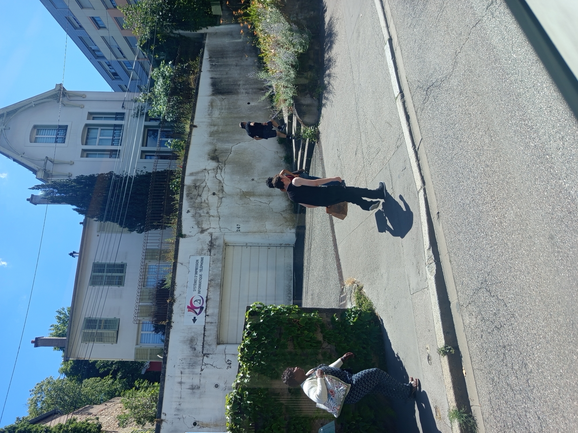 The image shows a street scene in Nancy, France, with several people walking and climbing stairs. In the background, there is a white building with green shutters, a tall building with a blue facade, and a large garage door with a sign that reads "SYSTÈMES D'IMPRESSION INFORMATIQUE - TÉLÉPHONIE". A woman in a patterned dress and white cardigan walks on the left side of the image, carrying a floral tote bag. Several people are walking up a set of concrete stairs on the right side of the image, with one person in a black t-shirt and shorts in the lead. The environment appears to be a sunny day with a clear blue sky.
