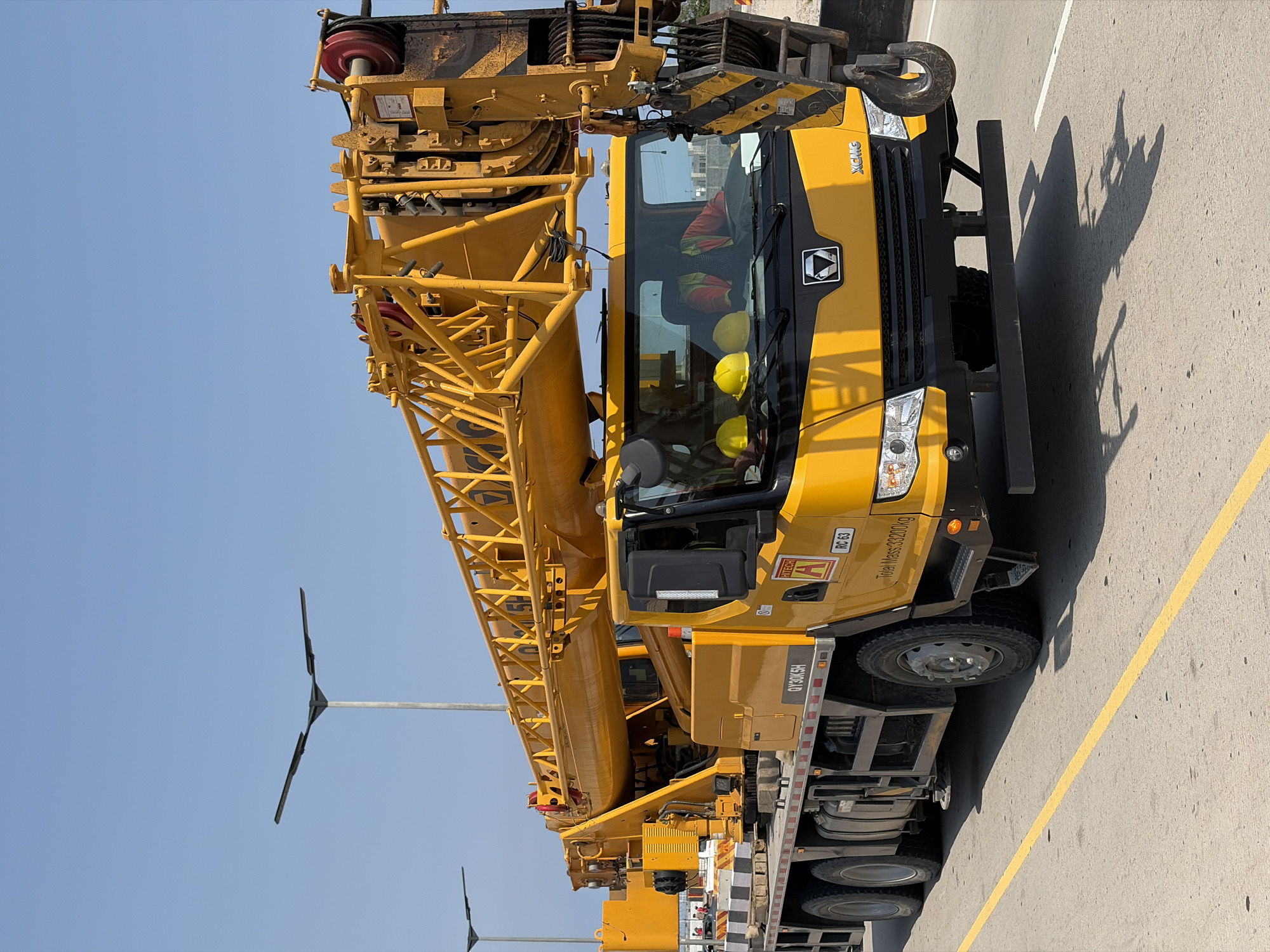 A large yellow XCMG crane is parked on an asphalt surface marked with a yellow line. The sky is clear and blue. The crane's boom extends toward the left. A few street lights are visible in the background. The location is Alaguntan, Nigeria. There are two hardhats inside the cabin. One is being worn by a person and the other is placed on the dash board. The crane model is QY30K5H. It has an "RC 63" sign on its side. "Total Mass: 33200 kg" is written on it.