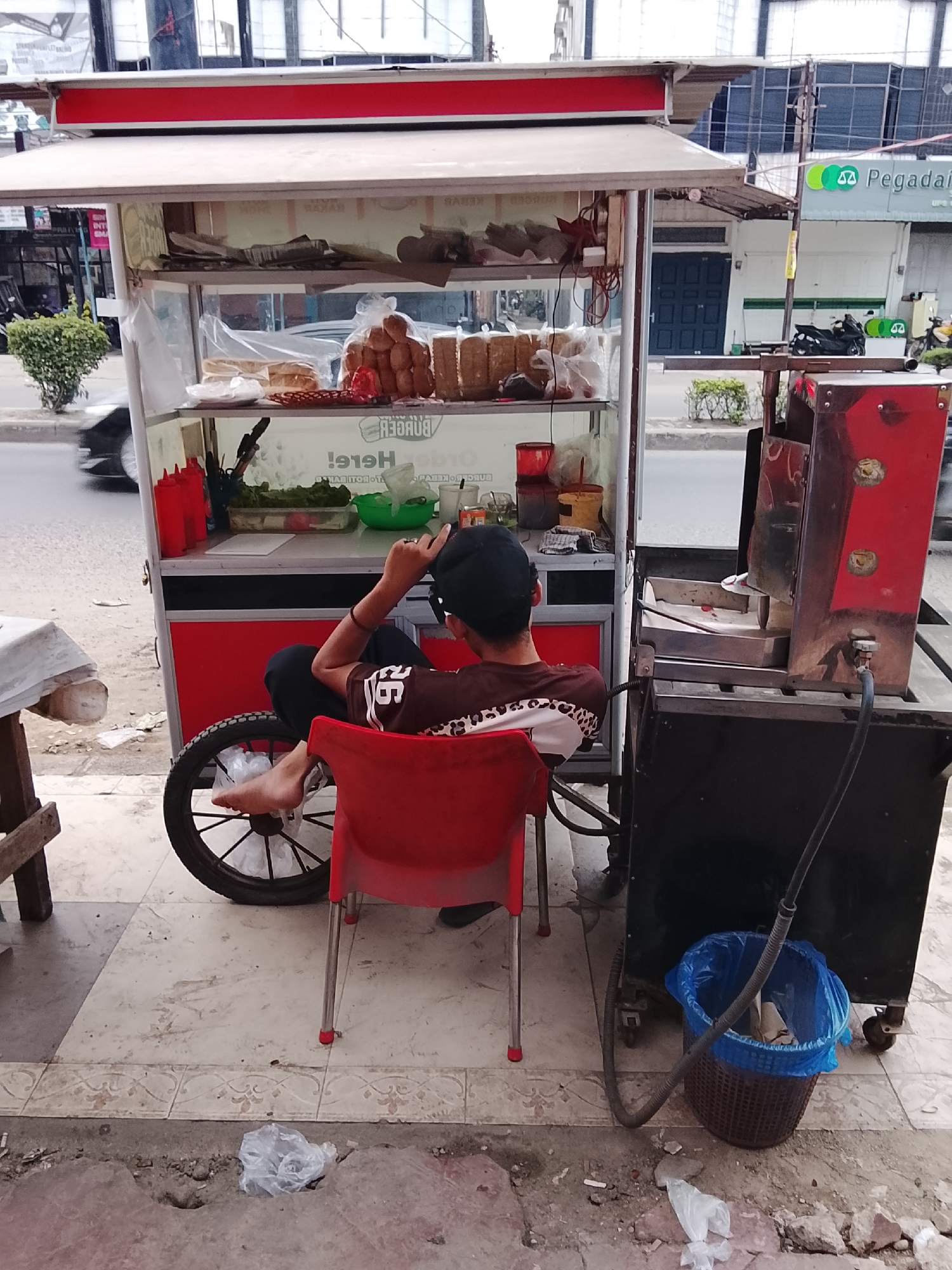In Lhokseumawe, Indonesia, a person sits in a red chair, facing away from the camera, at a mobile food cart. The cart is filled with various food items and condiments, including bread rolls, lettuce, and red sauce bottles. A large propane tank and a charcoal grill are attached to the side of the cart. The scene is set on a paved street with a sidewalk. The background shows buildings and a busy street with passing vehicles. The person is wearing a dark t-shirt and a black cap. The ground is littered with some trash.