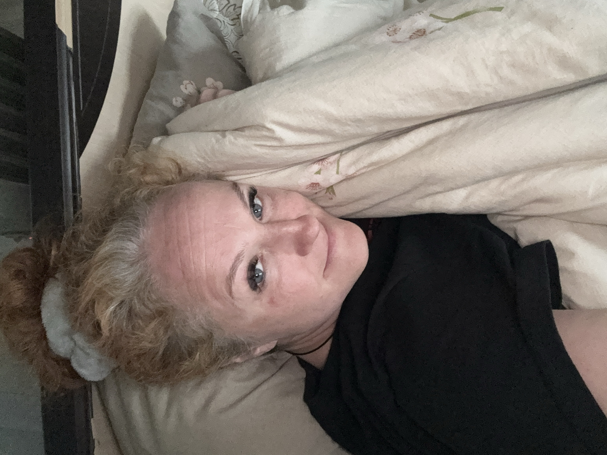 The image is a close-up, angled shot of a woman lying in bed. She has light skin, blue eyes, and curly, reddish-blonde hair pulled back in a bun with a light grey scrunchie. She is wearing a black shirt and a thin black necklace. She is looking directly at the camera with a slight smile. She is partially covered by a light-colored blanket with floral patterns. A patterned pillow is visible behind her. The headboard of the bed, made of dark wood, is visible in the upper left corner of the frame.
There is no information to suggest that the location is in the city of Chaikovsky, Russia.