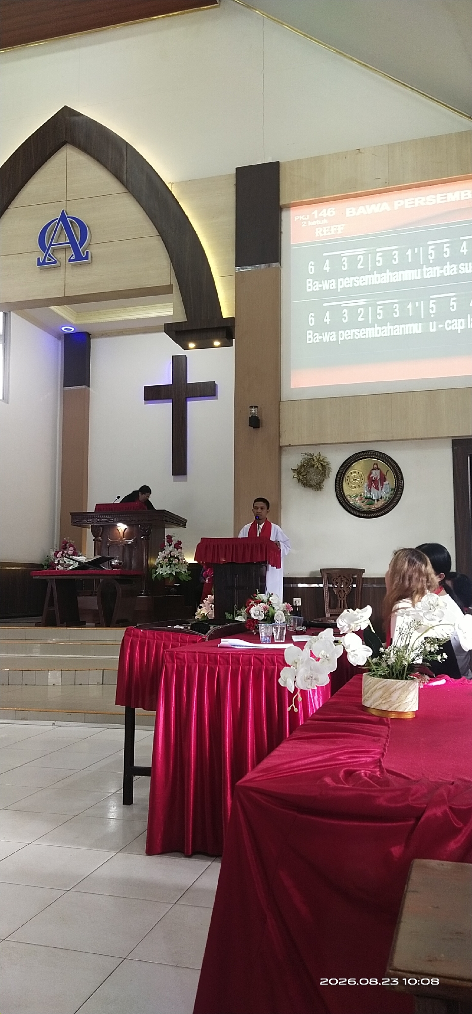 The image depicts the interior of a church, likely in Jaar, Indonesia. In the foreground, two tables are covered with vibrant red tablecloths, adorned with floral arrangements. One table holds two glasses and some papers. In the middle ground, a man in a white robe with a red stole stands behind a wooden podium, speaking to the congregation. To his left, another person is seated at a wooden pulpit, facing away from the camera. Above them, a large, dark wooden cross is mounted on the wall. The background features a projection screen displaying text in Indonesian, along with musical notes. To the left of the cross, a blue symbol resembling the Alpha and Omega is displayed above an arched entryway. The overall environment suggests a religious service is underway. The timestamp in the bottom right corner indicates the image was taken on August 23, 2026, at 10:08.