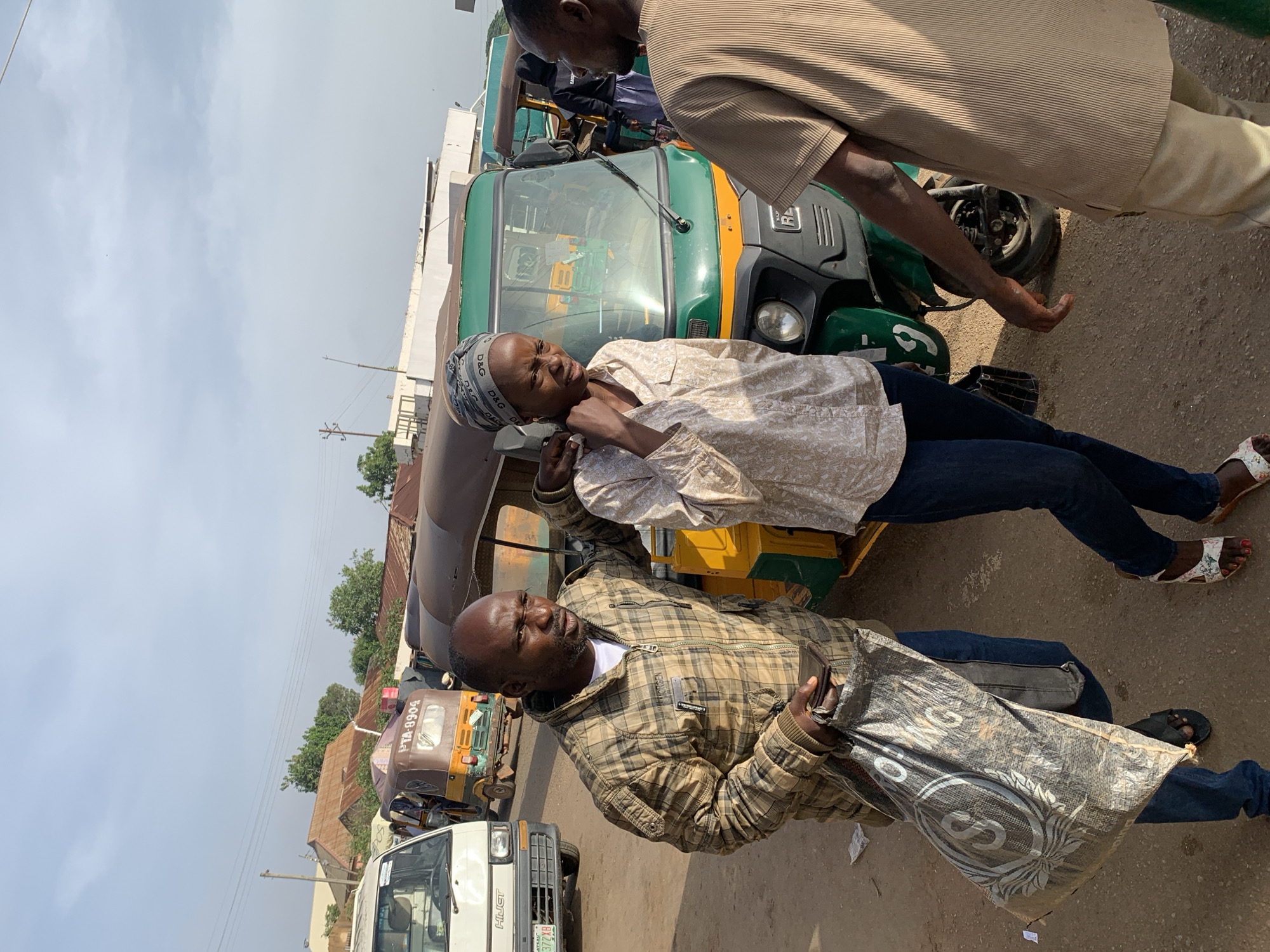The image depicts a street scene in Gura Top, Nigeria. Three individuals are the central focus, positioned in the foreground and mid-ground. A man on the left, wearing a brown plaid jacket and blue jeans, carries a large, dark plastic bag. He is looking towards the right of the frame with a stern expression. To his right, a woman, also in blue jeans and a light-colored patterned shirt, is being touched on the shoulder by the man. She is looking downwards and appears to be distressed or upset, with a cloth headwrap on. Another man, wearing a beige, ribbed shirt and light-colored trousers, is on the right side of the image, with his back to the camera, partially obscuring the woman.
In the background, several auto-rickshaws, commonly known as "Keke" in Nigeria, are parked or moving. One green and yellow auto-rickshaw is prominent behind the main subjects, with a visible license plate starting with "PTA". Other vehicles and buildings with corrugated metal roofs are visible further in the distance, suggesting a bustling urban or semi-urban environment. The ground is dusty and unpaved. The sky is overcast with a light blue hue. The overall impression is a moment captured in a busy public space, possibly a transport hub or market area.