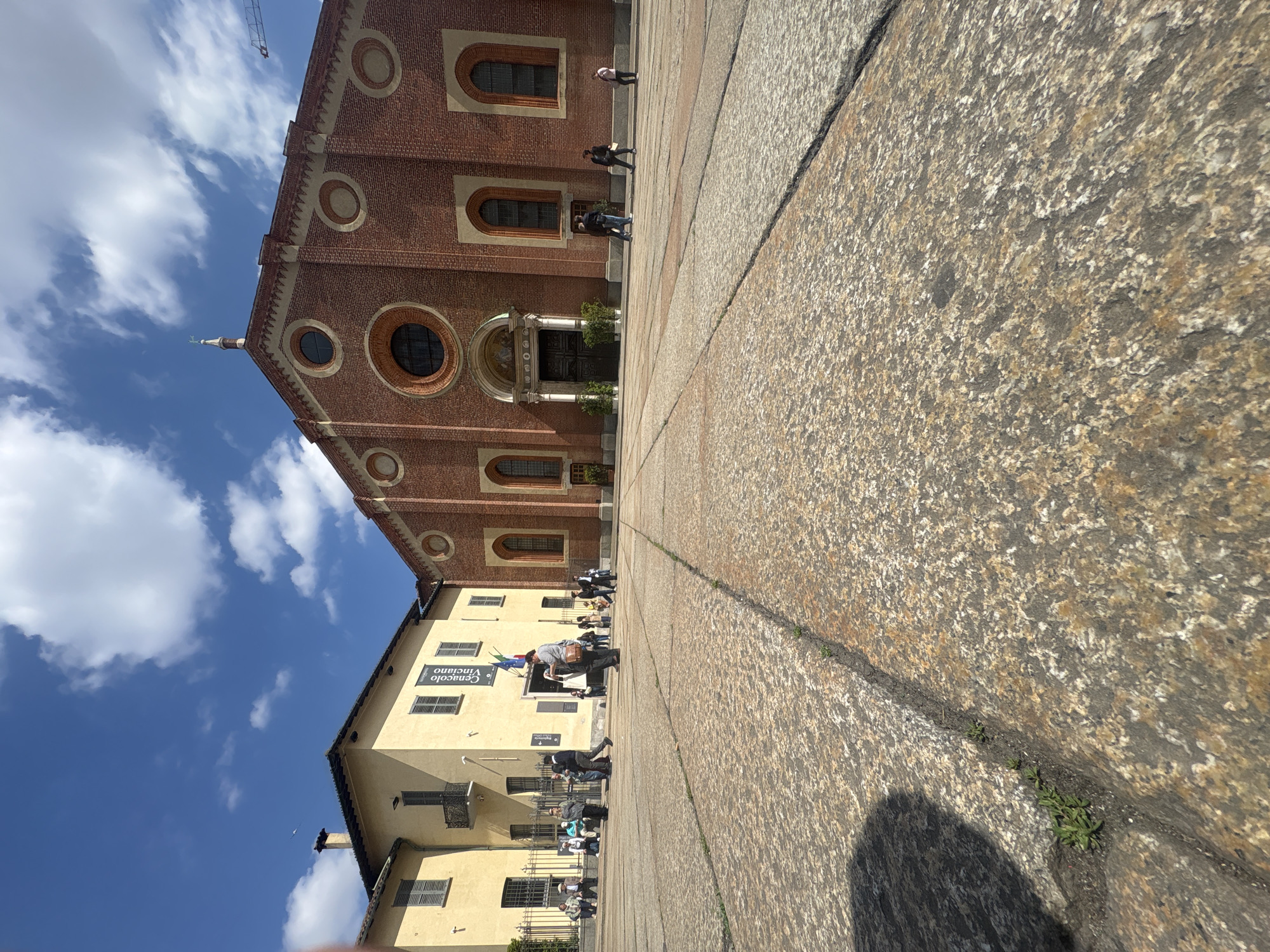 The image captures a scene in Milan, Italy, showcasing the Church of Santa Maria delle Grazie and a portion of the adjacent building, the Cenacolo Vinciano. The church, constructed of red brick, features rounded windows and a central doorway with a fresco above it. The Cenacolo Vinciano, with its pale yellow façade, displays signage indicating the entrance to the venue, likely where Leonardo da Vinci's "The Last Supper" is housed. Pedestrians populate the foreground and are interacting within the scene, suggesting this is a tourist location. The image has a low angle and is likely taken from the ground. The sky is a vibrant blue with scattered white clouds. The cobblestone pavement of the courtyard is also visible.