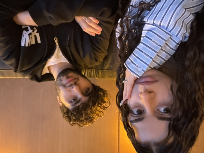 The image displays two individuals, a female and a male, seated indoors in Yerevan, Armenia.
The female, positioned on the left in the foreground, has dark, curly hair extending past her shoulders. She wears a light blue and white striped long-sleeved button-up shirt. Her left hand, featuring a ring on the ring finger and light-colored nail enhancements, rests against her left cheek and jawline. Her gaze is directed towards the right side of the frame, away from the camera, and her mouth is closed.
The male is seated to the right of and slightly behind the female. He has dark, curly hair and a short beard. He is wearing a black zip-up hoodie, which features a white "LAVA" logo on the left chest area, over a white t-shirt. A ring is visible on his left ring finger, and his fingernails are painted a dark color. His arms are crossed over his chest, and he looks directly at the camera with a subtle smile, his head tilted slightly to the viewer's right.
Both individuals are seated on a dark-colored, possibly leather or faux-leather, cushioned surface. The background consists of a plain, warm brown wall. The scene is illuminated by dim, warm, and indirect lighting. The specific establishment or precise address within Yerevan, Armenia, is not discernible from the visual information provided.