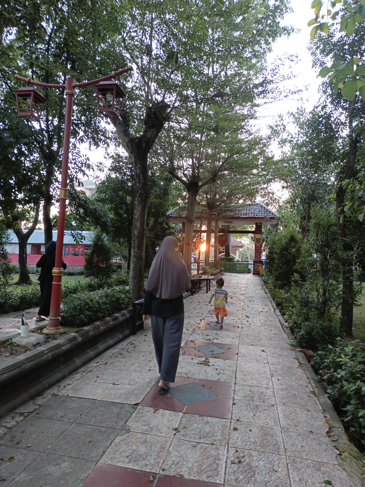 In a park setting in Klaten, Indonesia, a paved walkway is bordered by lush green trees and bushes. The walkway is made of large square tiles, some of which have diamond-shaped inlays of red and gray. Fallen leaves are scattered across the path. A decorative red lamppost with two lantern-like fixtures stands to the left of the frame. In the background, a traditional-style pavilion with a red-tiled roof is visible, its entrance framed by decorative posts. A street lamp with an orange glow is visible through the pavilion's archway.
Two individuals are walking away from the camera. The person in the foreground, wearing a grey hijab and dark green long-sleeved top with dark grey pants, walks on the right side of the path. To the left and further down the path, a child in a striped t-shirt and orange shorts walks ahead. Another person wearing a black hijab and black clothing is partially visible on the far left of the image, near the base of the lamppost. The scene appears to be during dusk, with soft lighting and shadows suggesting the sun is setting.