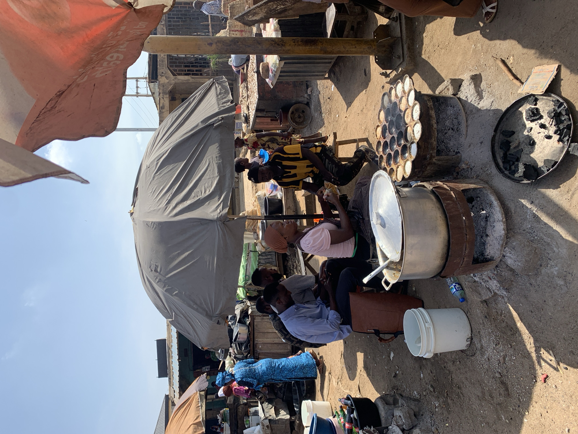 **Location:** Rafin Par, Nigeria.
**Scene:** An outdoor, likely makeshift, cooking and market area. Several people are seated around what appear to be cooking stations.
**People:**
* Several individuals are seated, some appear to be eating, while others are likely involved in the cooking or selling process.
* Clothing styles vary; some wear casual attire, while others wear more traditional clothing. Headscarves are visible.
**Objects:**
* Large metal pots and pans, some used for cooking over charcoal stoves made from modified barrels.
* A charcoal grill sits beside a charcoal pot filled with charcoal.
* Utensils for stirring food.
* Plastic buckets and bottled drinks.
* Umbrellas providing shade.
* A stove with circular holes for cooking.
**Actions:**
* Cooking and food preparation are occurring.
* Eating is taking place around the cooking area.
* Likely, selling and purchasing of food are happening.
**Interactions:**
* People are interacting and seated around the cooking stations.
**Environment:**
* A crowded, dusty outdoor area, suggesting a market or street scene.
* Various stalls and makeshift structures are visible in the background.
* Partial shade is provided by umbrellas.