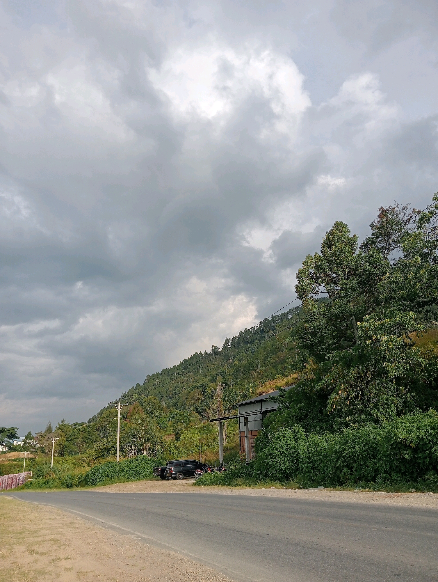 The image depicts a roadside view in the city of Hinalangbagasan, Indonesia. The foreground shows a paved asphalt road curving to the left, with a gravel shoulder on its left side. On the right side of the road, a dense growth of green vegetation lines the edge, obscuring a portion of the scene behind it. A dark-colored SUV is parked on the side of the road, partially visible behind the vegetation. Further back, a small, simple structure with a metal roof and wooden walls is nestled amongst the greenery.
Behind the roadside and vegetation, a steep, tree-covered hill dominates the background. The hillside is densely populated with a variety of green trees, with some electrical poles and wires running along its slope. The sky above is overcast, filled with heavy, grey clouds, suggesting a possibly stormy or cloudy day. In the far distance, on the left side of the image, a few buildings and structures can be seen at the base of another, less steep incline. The overall scene conveys a rural or semi-rural setting with natural elements and infrastructure present.