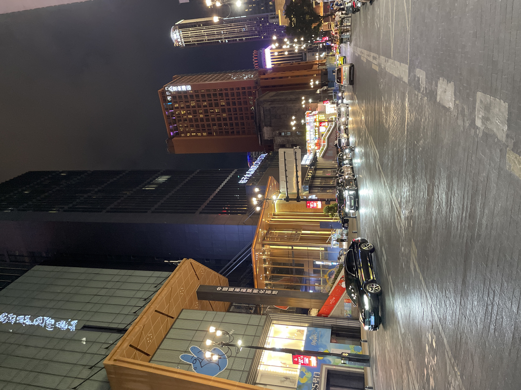 The image depicts a street scene at night in the Yuzhong District of China, characterized by illuminated buildings, streetlights, and moving vehicles. On the left, a modern building with a glass facade displays blue neon text. Adjacent to it is a structure with an orange awning and a blue graphic of a hand with two fingers raised. A red staircase leads upwards between these buildings. In the foreground, a black sedan is driving on a wet, cobblestone-like street.
Further down the street, several other vehicles are visible, including cars and what appears to be a motorcycle. The street is lined with tall buildings, many of which are illuminated with various colored lights. One prominent building is orange and displays illuminated Chinese characters. To its right, a taller, slender building is also lit up. Numerous streetlights with multiple bulbs illuminate the road. The sky above is dark, suggesting it is nighttime. A few pedestrians can be seen on the sidewalks and near the buildings. The overall atmosphere is urban and bustling.
