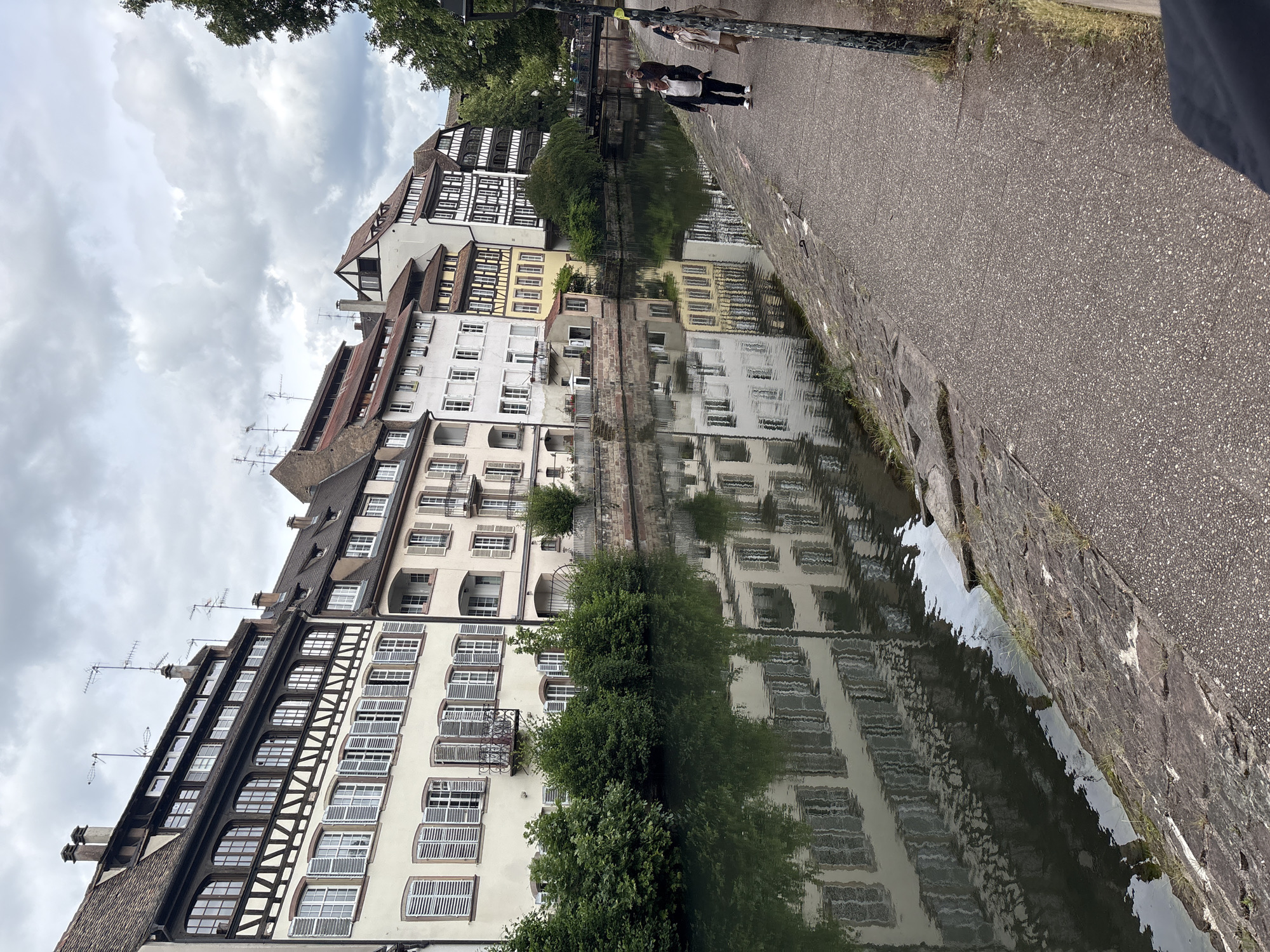 The image captures a picturesque canal in Strasbourg, France, specifically in the Petite France district. The scene features a row of traditional Alsatian half-timbered houses lining the canal, their facades reflected in the calm water. The architecture is characterized by dark wooden beams contrasting with pale stucco walls and numerous windows with shutters. Lush green trees and bushes grow along the canal bank, adding a vibrant natural element to the urban landscape. A paved walkway runs alongside the canal, and several individuals are seen strolling along it. One person in the foreground is partially visible, wearing dark clothing. Further down the path, two individuals are walking together, one in a white jacket and dark pants, and the other in a light-colored coat. In the distance, a bridge crosses the canal, and more buildings are visible on the opposite bank. The sky is overcast with clouds, casting a soft, diffused light on the scene. The overall impression is one of a charming and historic European city.