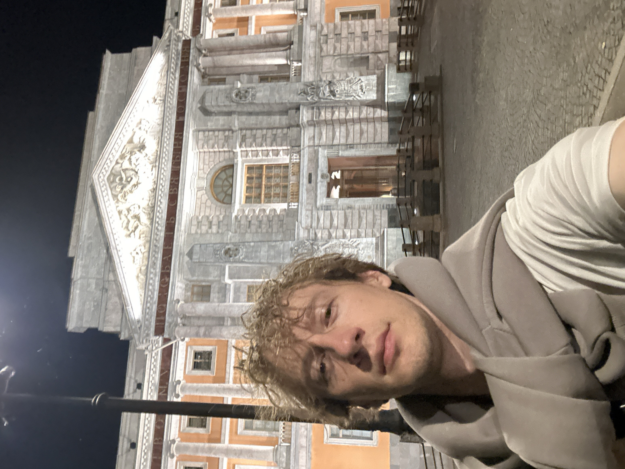A man with curly blonde hair takes a selfie at night in front of a large, ornate building in Saint Petersburg, Russia. The building, illuminated by lights, features classical architectural elements such as columns, intricate carvings, and a triangular pediment. The text "БЫТЬ СВЯТЫНЯ ГОСТИНА" is visible above the entrance. The man is wearing a grey hoodie draped around his shoulders over a white t-shirt. The cobblestone ground and a lamppost are also visible in the frame.