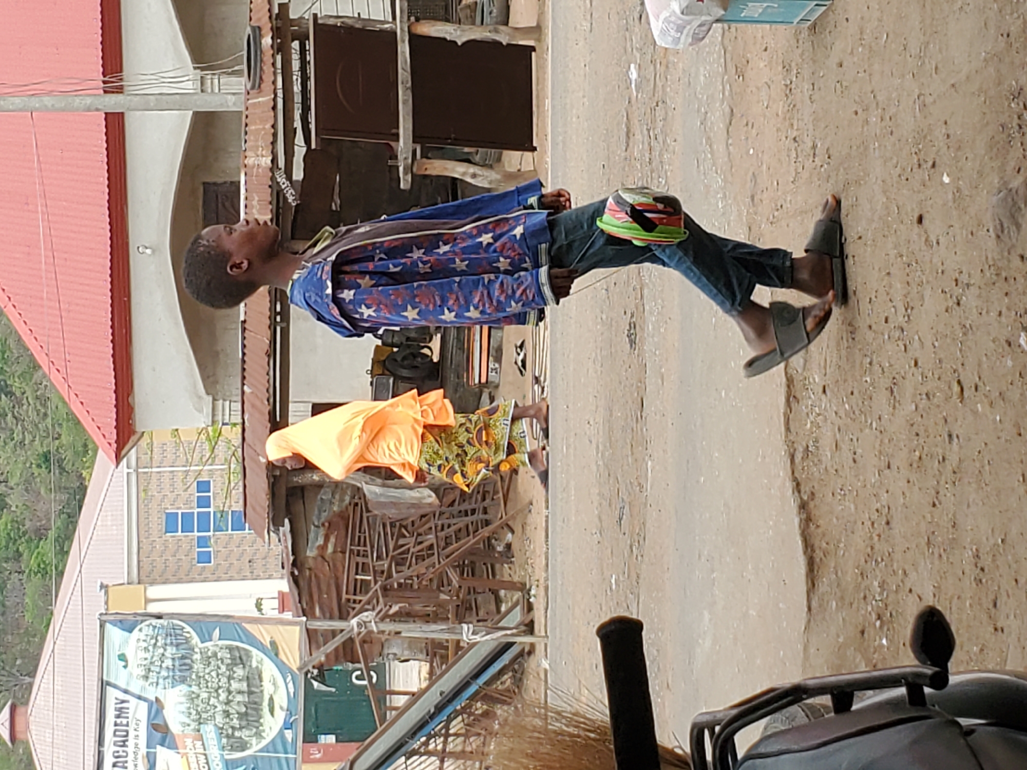 A young Black male stands on a dusty, possibly unpaved street in Mararaba, Nigeria. He wears a blue jacket with white stars, dark jeans, and gray sandals. A colorful helmet is positioned on his right knee. He gazes upwards. In the background, buildings with corrugated iron roofs are visible, including one with a red roof and another with a blue cross design on its exterior. A banner advertises an "ACADEMY" with the slogan "Knowledge is Key". Debris and metal scaffolding are scattered near a figure in an orange cloth. Part of a motorcycle or quad bike is visible in the foreground.