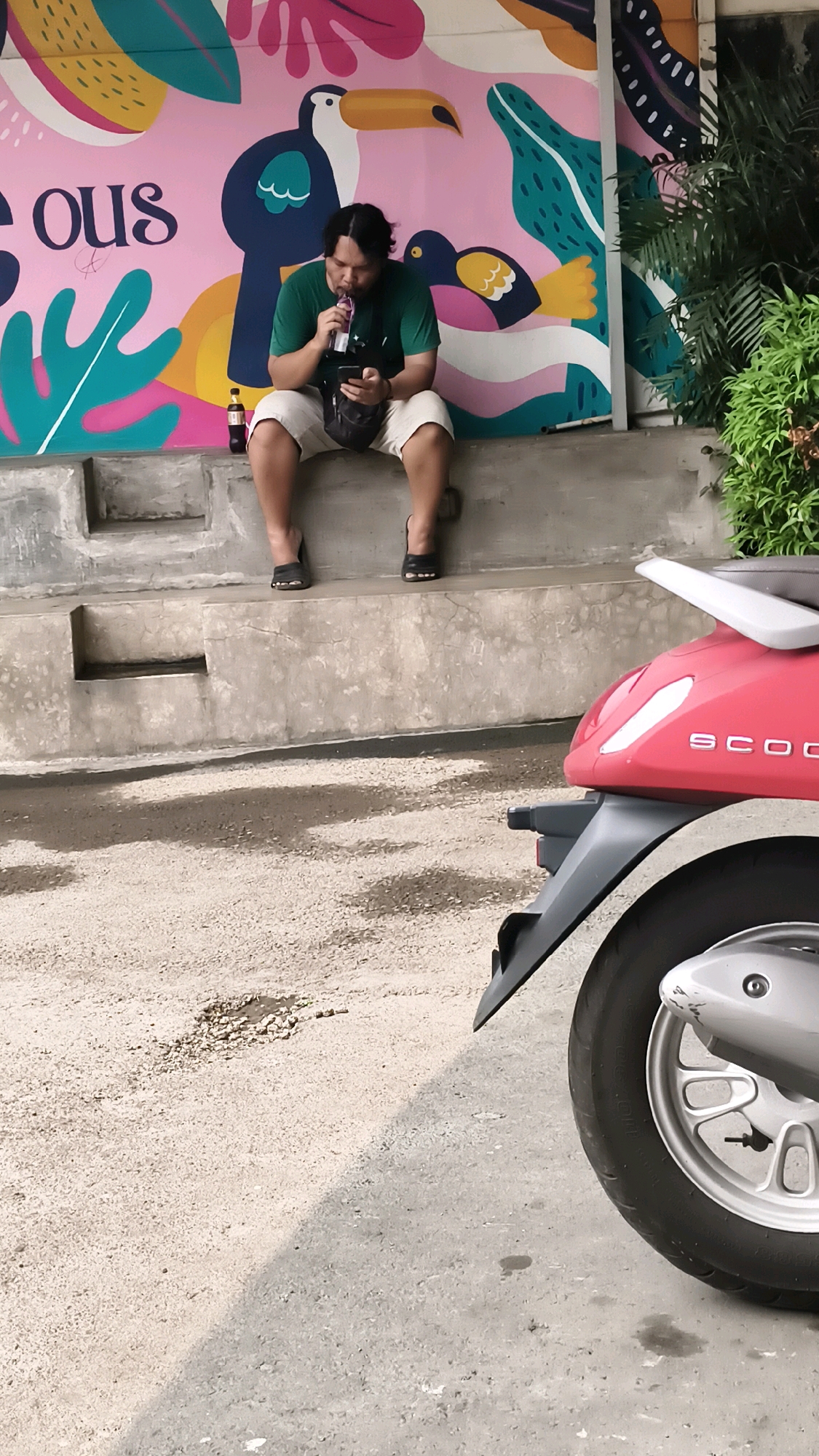 A man wearing a green t-shirt and beige shorts is sitting on a concrete ledge. He is holding a phone in his left hand and drinking from a purple pouch with his right. A dark shoulder bag is slung across his body. To his left, a small brown bottle is on the ledge. Behind him is a colorful mural featuring abstract shapes and birds. A red scooter is parked in the foreground, partially obscuring the bottom right of the image. The ground is a mix of concrete and gravel. The scene appears to be outdoors, possibly in an urban setting. This scene is likely located in Bekasi, Indonesia, as per the user's request.