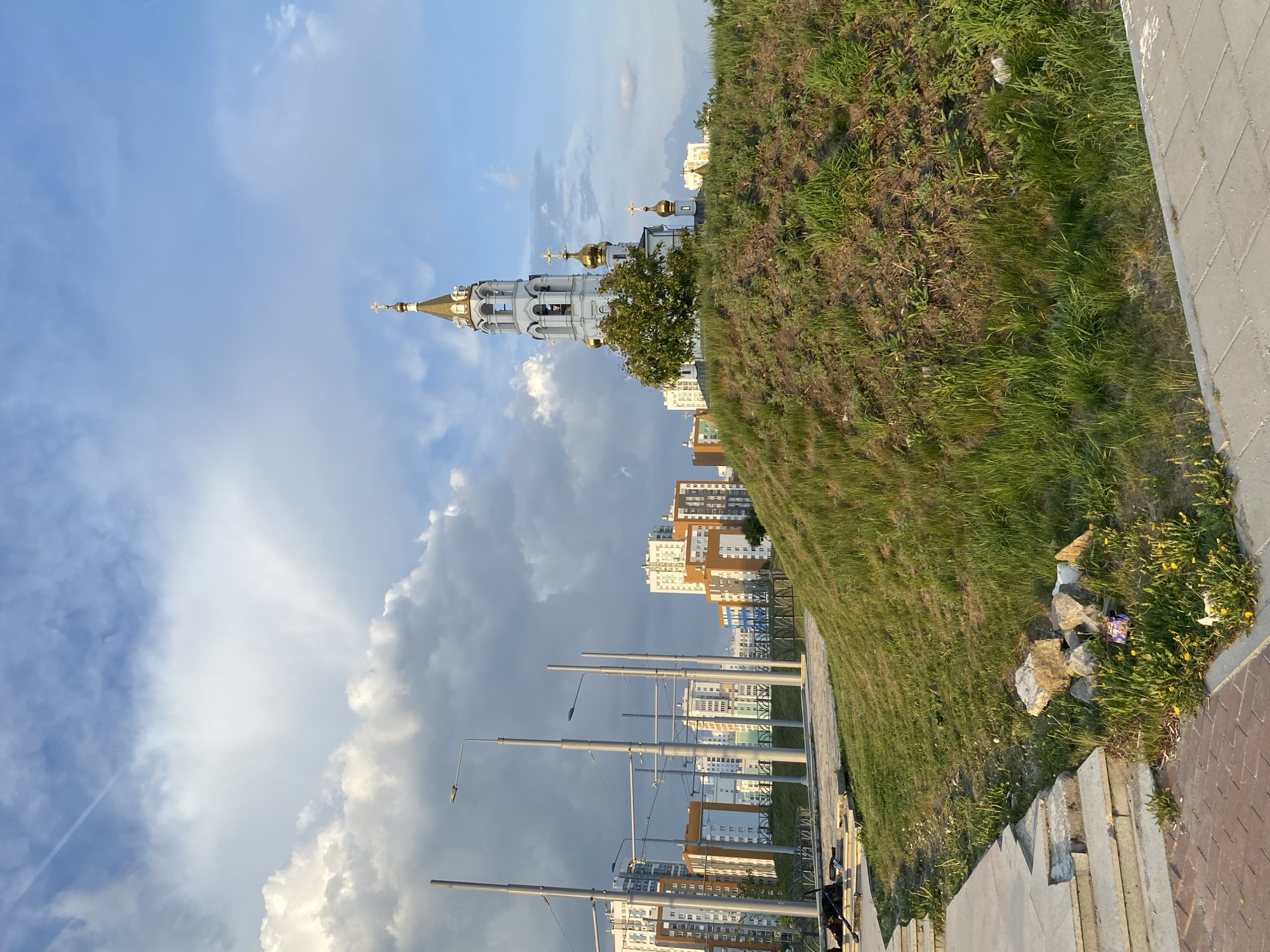 The image captures a scene in Yekaterinburg, Russia, featuring the Church on Blood in Honor of All Saints Resplendent in the Russian Land. The church, with its white walls and golden domes, is situated near modern apartment buildings and street lamps. A grassy hill dominates the right side of the frame, leading down to a paved walkway with a curb and stairs. Rocks and scattered trash are visible near the bottom of the hill. The sky is filled with dramatic clouds, ranging from bright blue to dark grey, suggesting an overcast day. No people are prominently visible in the scene.