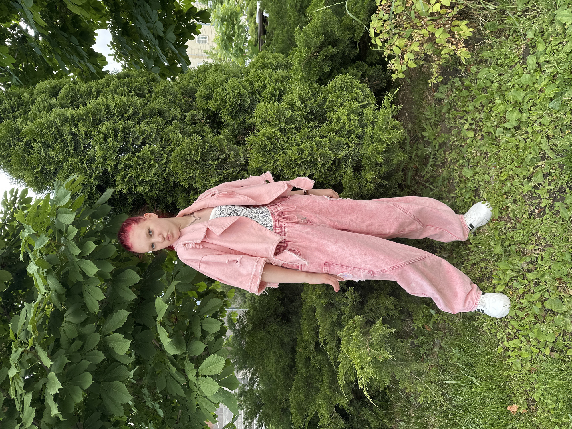 A young woman with red hair is lying on a patch of grass surrounded by trees and shrubs. She is wearing a pink denim jacket and pants, with white sneakers. The location appears to be a green space within a city, possibly a park or garden, given the variety of trees and maintained grass. The photograph was taken outdoors during the daytime. This photo was taken in Saratov, Russia.