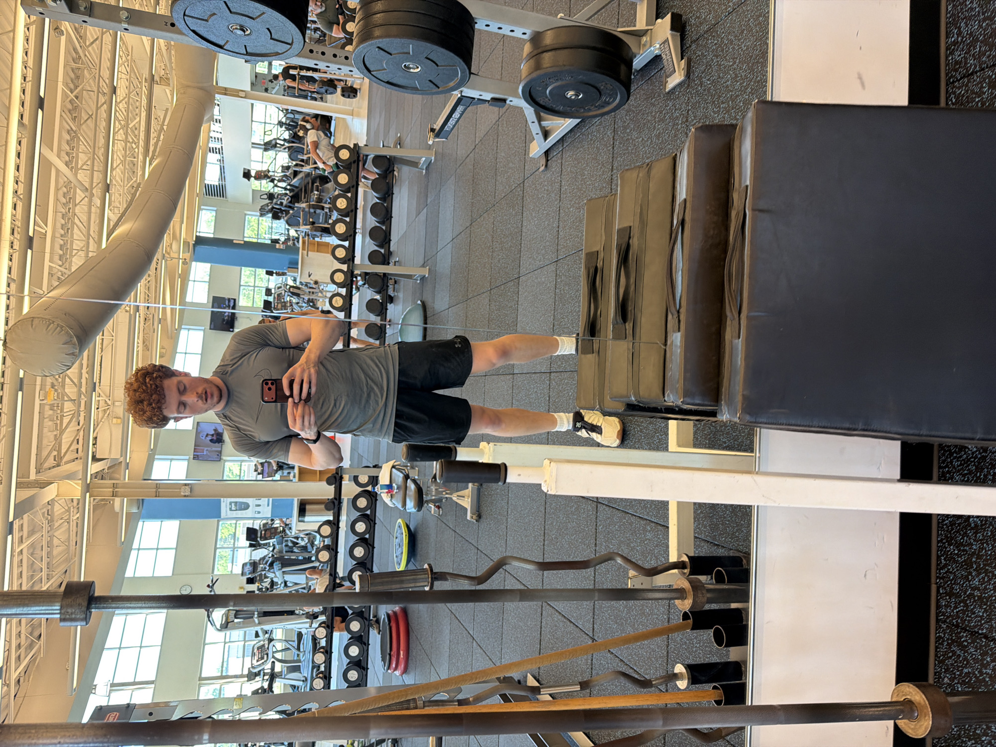 A man with curly red hair stands in a gym, taking a mirror selfie. He is wearing a gray t-shirt and black shorts. In the background, various gym equipment is visible, including weight racks, treadmills, and barbells. The gym appears to be well-lit and spacious.