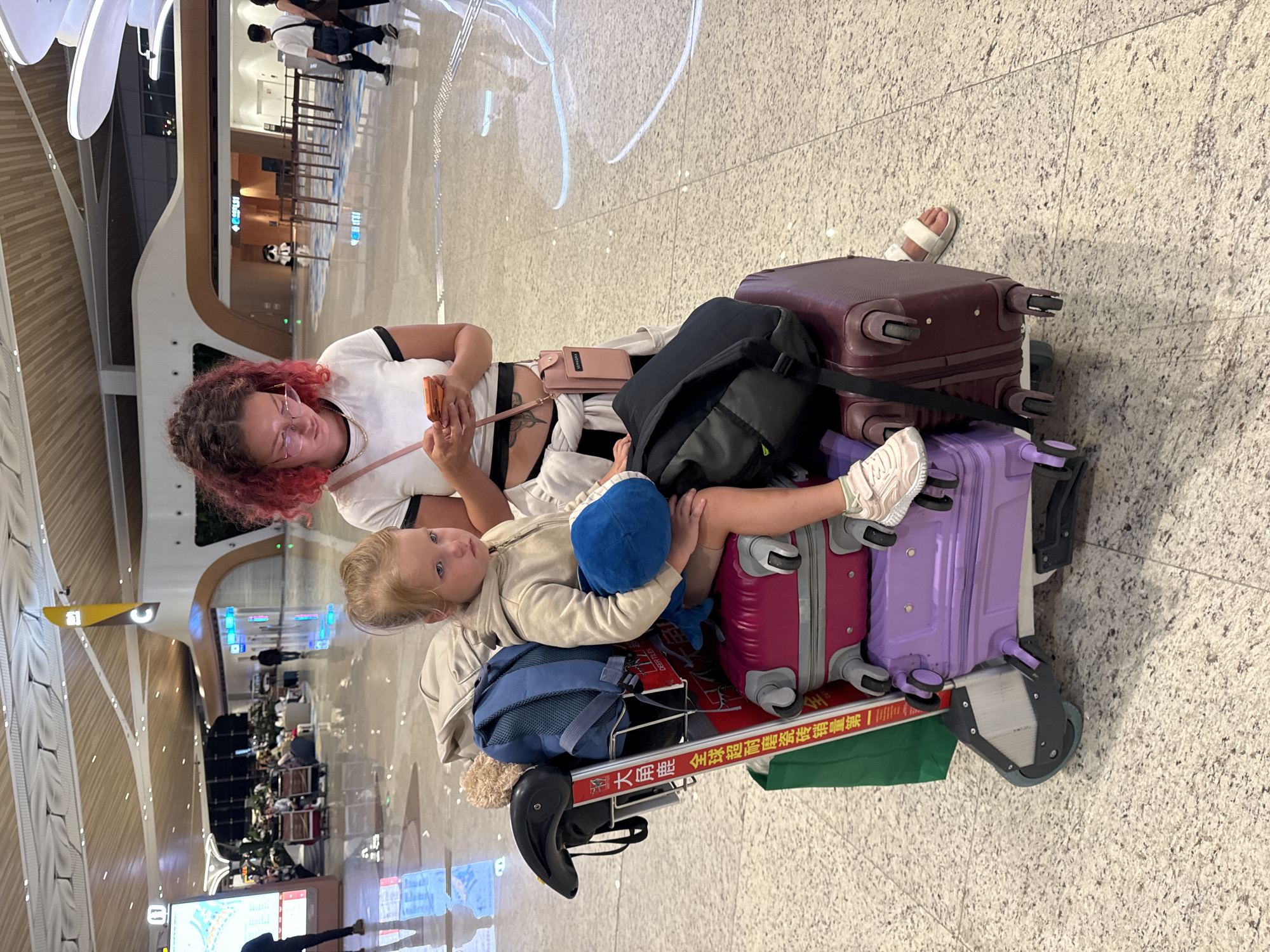 In an airport terminal, likely Baiyun District, China, a young girl with blonde hair sits on a luggage cart laden with several suitcases. She is holding a blue stuffed toy and looking down with a solemn expression. A woman with vibrant pink and red curly hair, wearing glasses and a white t-shirt, stands behind the girl, engrossed in her smartphone. The luggage cart is filled with various bags and suitcases, including a pink suitcase, a purple suitcase, and a dark red suitcase. A green shopping bag hangs from the side of the cart. The floor is a polished speckled tile. In the background, other travelers can be seen walking and sitting, and the airport's modern architecture with illuminated ceiling panels and decorative elements is visible.