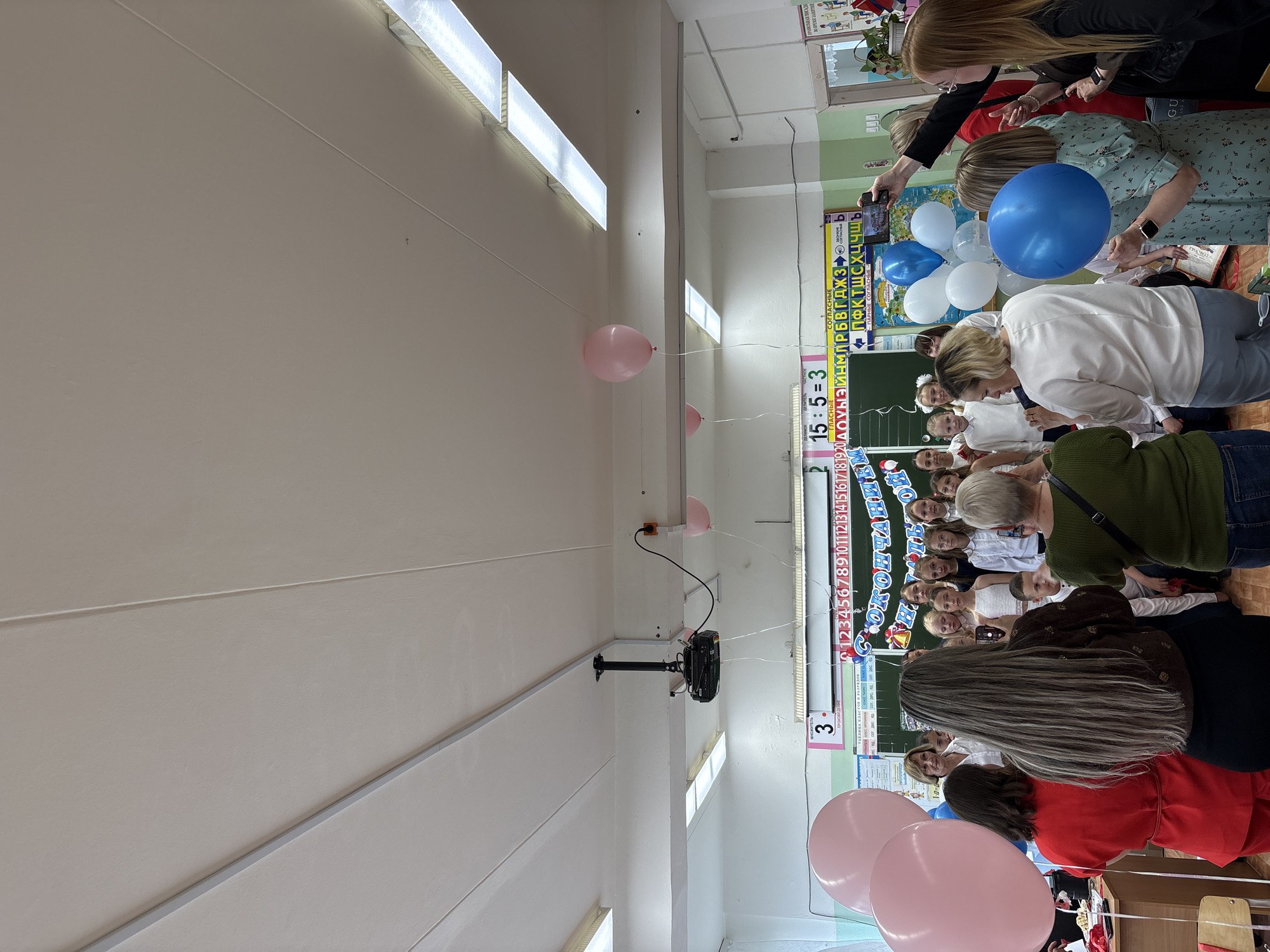 The image shows a gathering of people, mostly women, in what appears to be a classroom. Some of them are holding balloons in colors of pink, white, and blue. The room has fluorescent lights and a chalkboard with Russian writing and numbers. A projector is mounted on the ceiling. The location is suggested to be Chaikovsky, Russia, based on the Cyrillic writing.