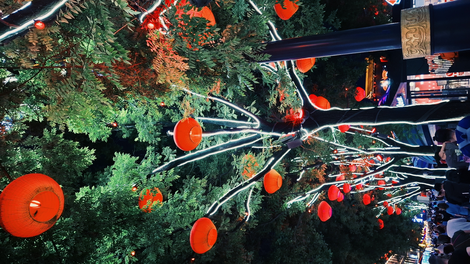 Yanta District, China: An outdoor nighttime scene shows trees illuminated with white LED light strands and red Chinese lanterns. The trees line a pedestrian area crowded with people. A black metal beam with bronze-colored decorative details spans across the frame. Buildings with glowing signs are visible in the background.