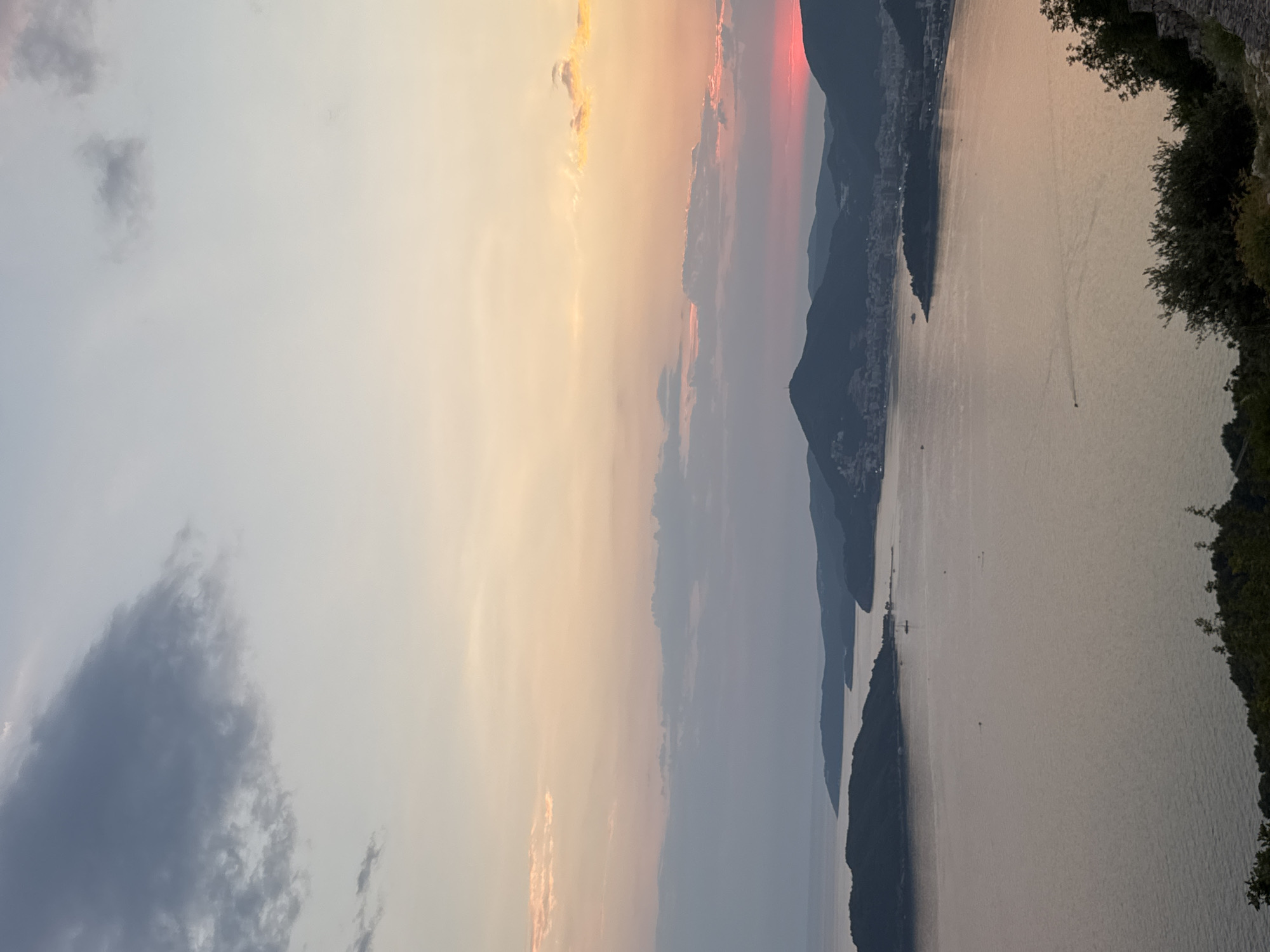 This is a high-angle, wide shot of a coastal bay at sunset in Kamenovo, Montenegro. The sky transitions from a pale blue and grey at the top to warm orange and pink hues closer to the horizon, where the sun is setting behind silhouetted mountains. A large island with a town is visible on the left side of the bay, connected to the mainland by a thin strip of land. On the right side of the bay, a larger mountainous coastline is densely populated with buildings. The water in the bay is calm and reflects the colors of the sky. Several small boats are scattered across the water. The foreground features lush green trees and rocky terrain.
