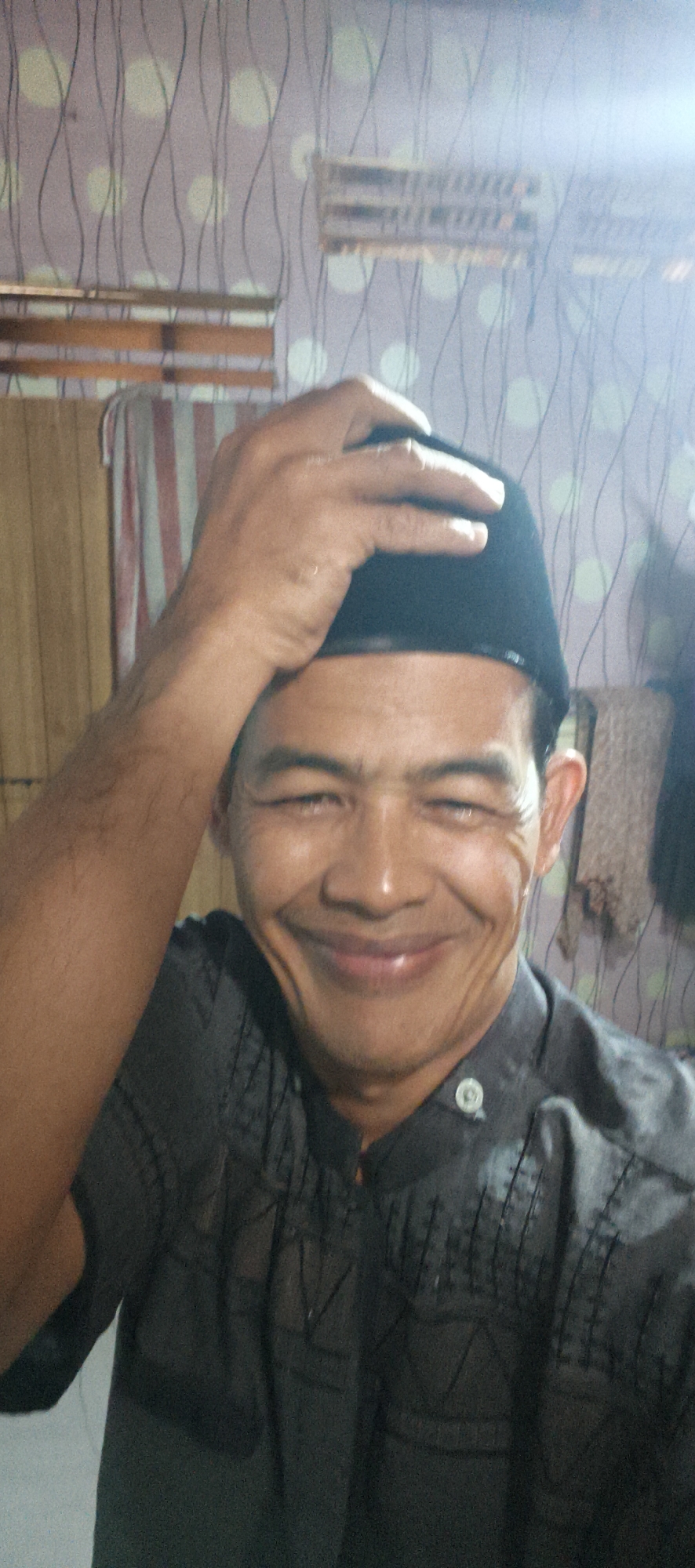 A close-up, waist-up, slightly angled, medium shot shows a middle-aged Indonesian man with brown skin, dark hair, and a smiling expression, wearing a black kufi and a dark gray, patterned button-up shirt. He is standing in front of a wall with a light purple patterned wallpaper, with vertical black lines and circular, lighter shapes. To the left of the man is a wooden shelf with what appear to be some textiles or hangings. The lighting is bright, with some overexposure on the right side of the frame. The overall impression is a casual, indoor portrait. The location is specified as Muaro Jambi, Indonesia.